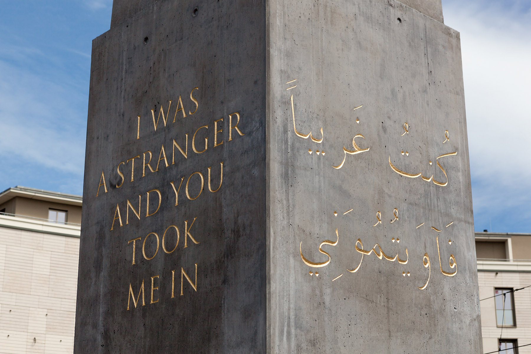Concrete pillar outdoors with the gold inscription “I was a stranger and you took me in” in English and Arabic script on the other side; buildings and a cloudy sky are visible in the background.