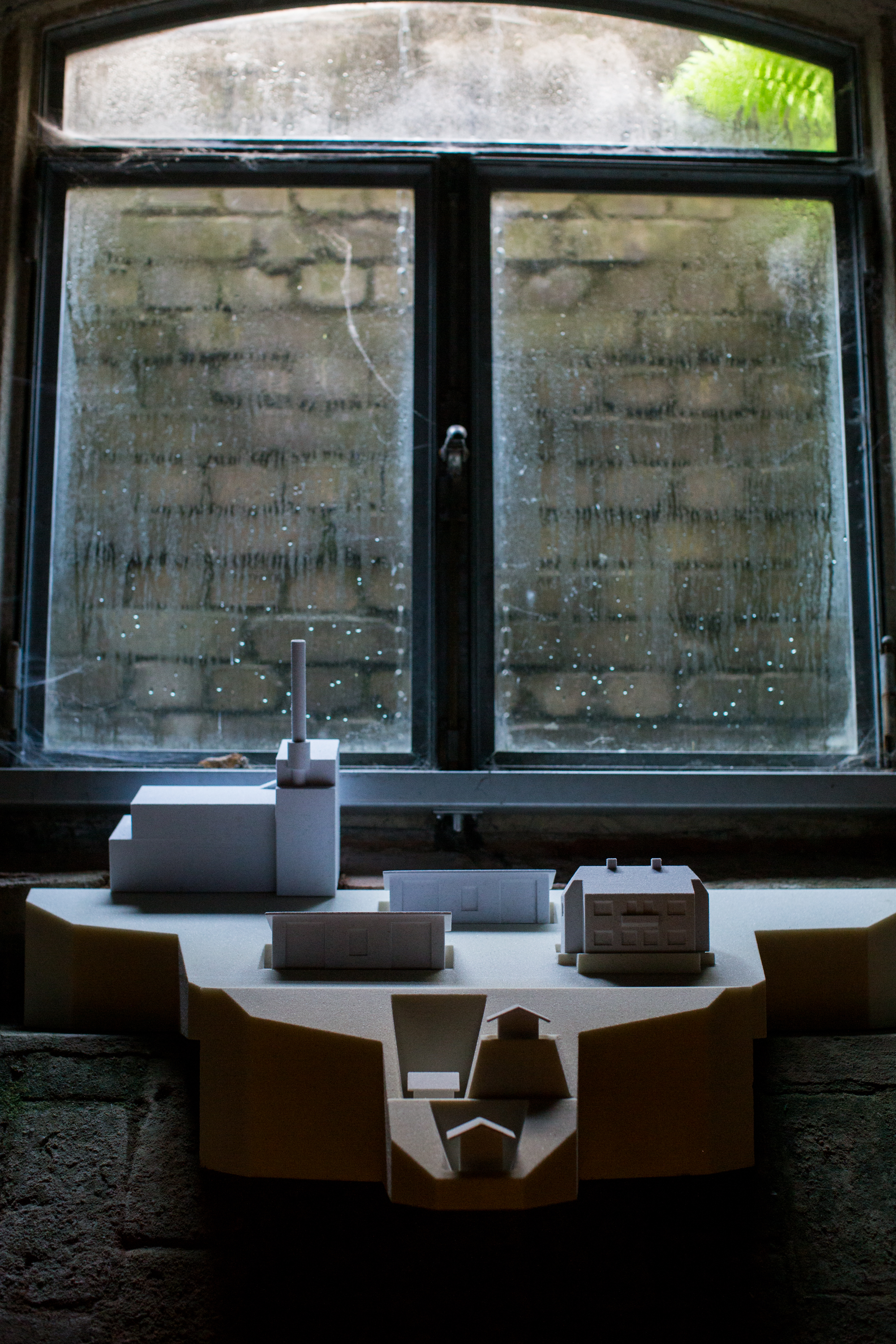 Architekturmodell einer Industrieanlage mit Wohngebäuden auf einer Fensterbank in einem historischen Gebäude. Das weiße Modell steht vor einem beschlagenen Fenster und kontrastiert mit der verwitterten Architektur und dem gedämpften Licht des Raumes.