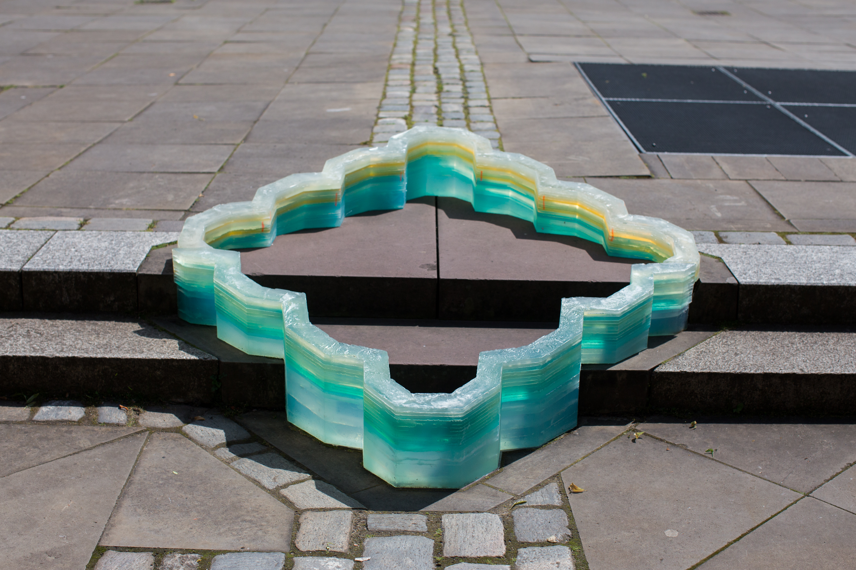Transluzente, türkisfarbene Skulptur aus geschichteten Materiallagen, die ringförmig über mehrere Stufen eines gepflasterten Platzes verläuft und eine geometrische Form bildet.