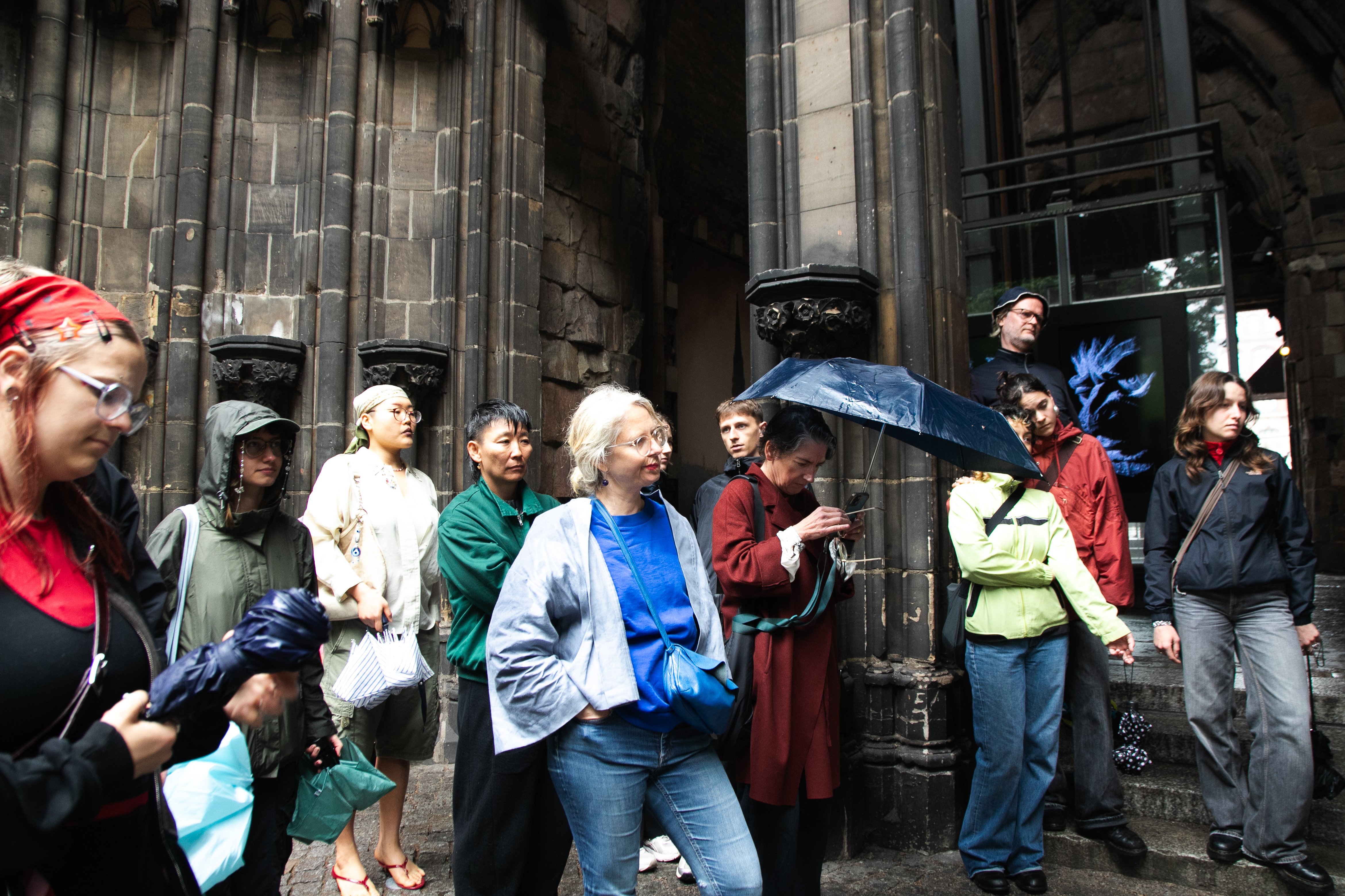 Eine Besucher*innengruppe steht im Eingangsbereich des Mahnmals St. Nikolai und verfolgt aufmerksam eine Führung oder Performance. Einige tragen Regenschirme oder Regenjacken, während sie vor den dunklen gotischen Säulen des Bauwerks versammelt sind.