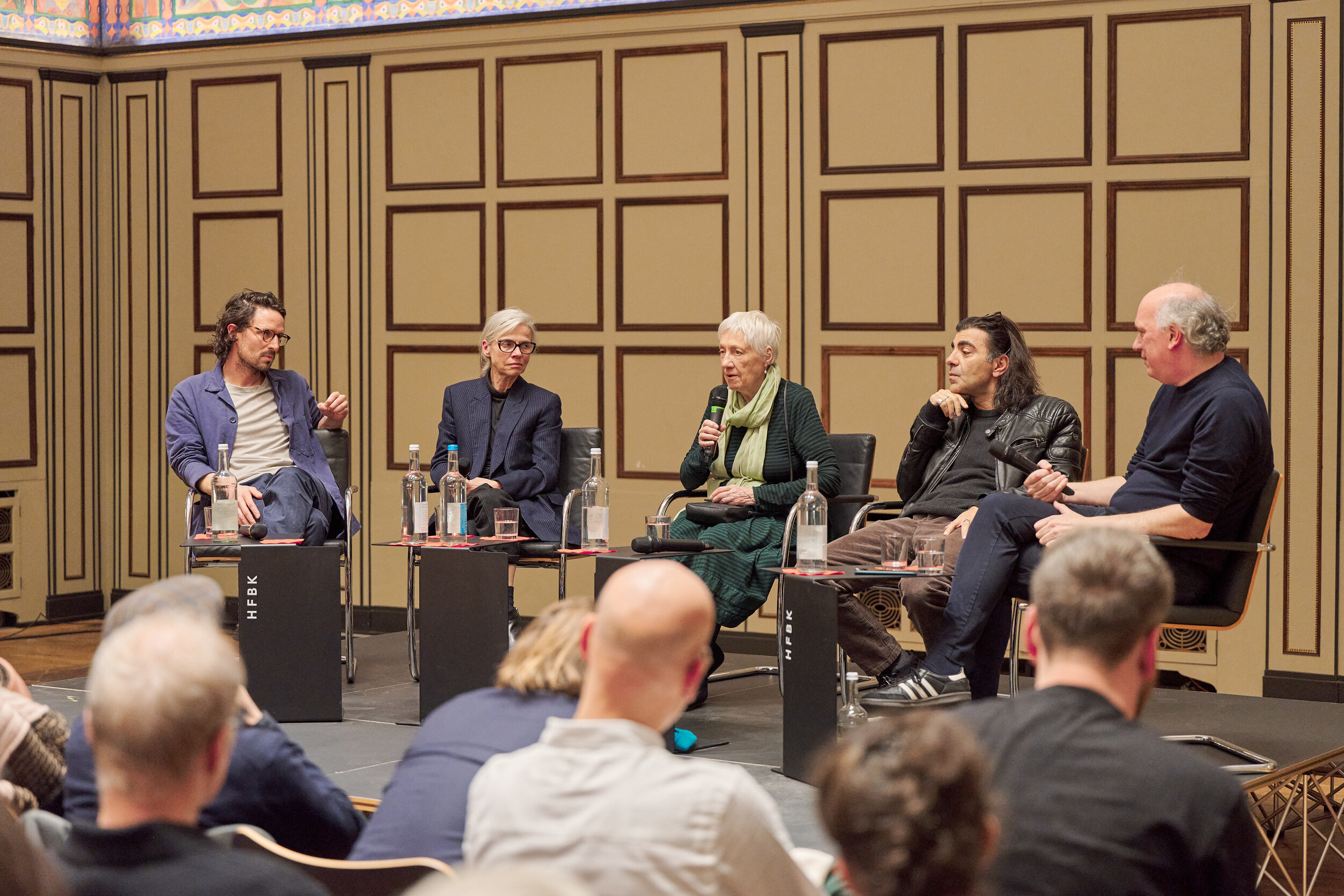 Five people sit on a stage in a wood-paneled hall during a panel discussion; an older woman speaks into a microphone, small tables with water bottles stand in front of them, and the audience is visible in the foreground.