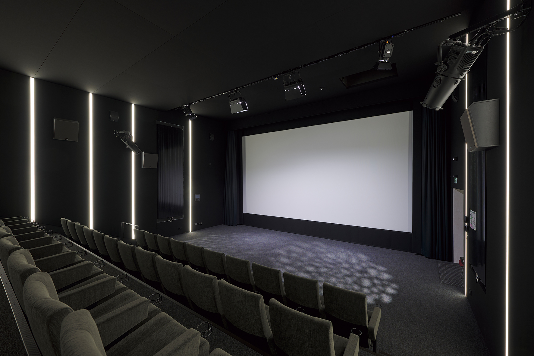 A modern cinema auditorium with several rows of cushioned seats, a large screen at the front, and vertical light strips on the dark walls.
