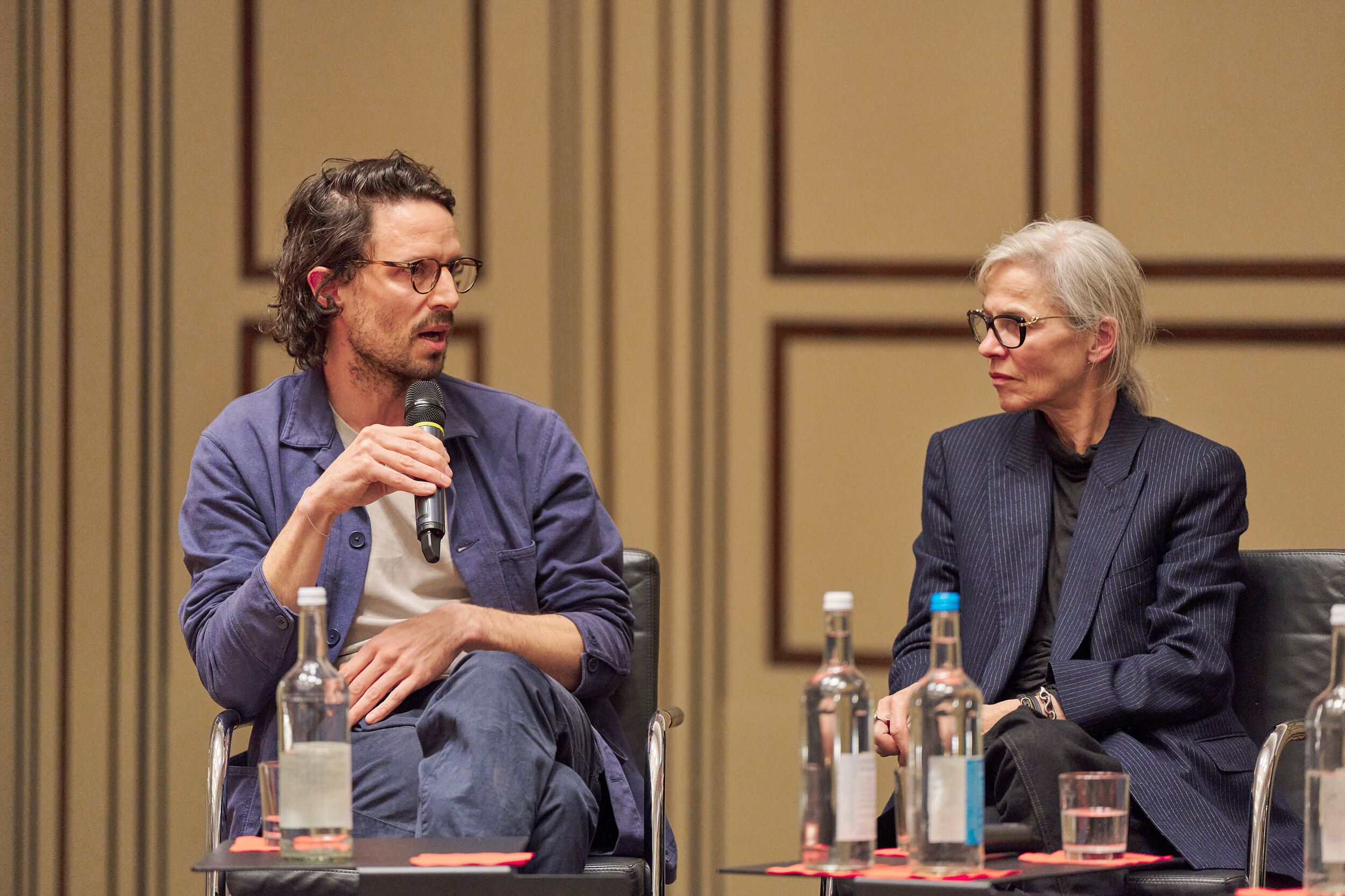 Two people sit on a stage in front of a wood-paneled wall, engaged in conversation. The man holds a microphone and speaks while the woman listens attentively. Several water bottles and glasses are placed on a table in front of them.