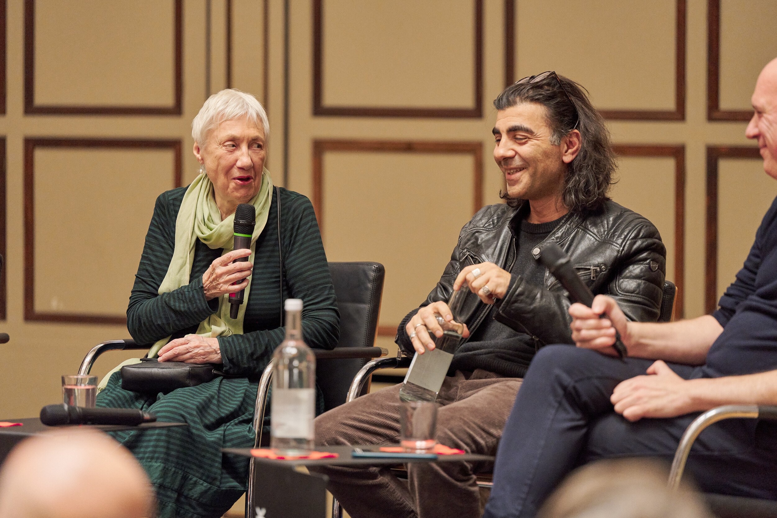 Panel discussion in a hall with wood-paneled walls. An elderly woman with short white hair and a green scarf holds a microphone while speaking. Next to her, a man with shoulder-length dark hair and a leather jacket smiles and holds a glass bottle. Another man sits to the right holding a microphone. Water bottles and glasses are placed on a table in front of them.