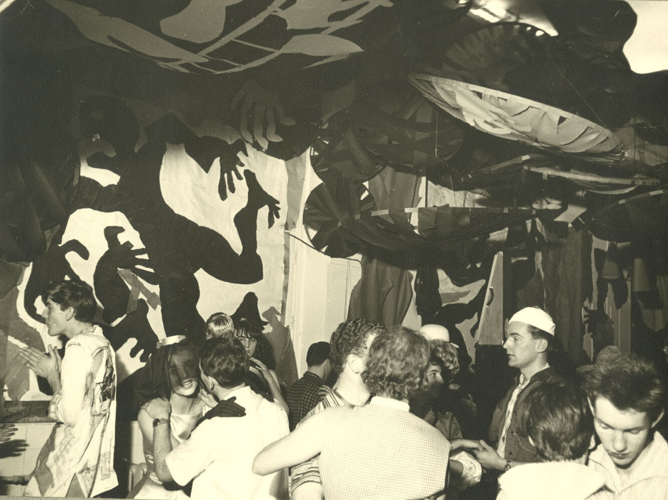 Historic black-and-white photograph of a party or exhibition scene: Several young people stand and dance in a room featuring large, high-contrast wall paintings and hanging decorations.