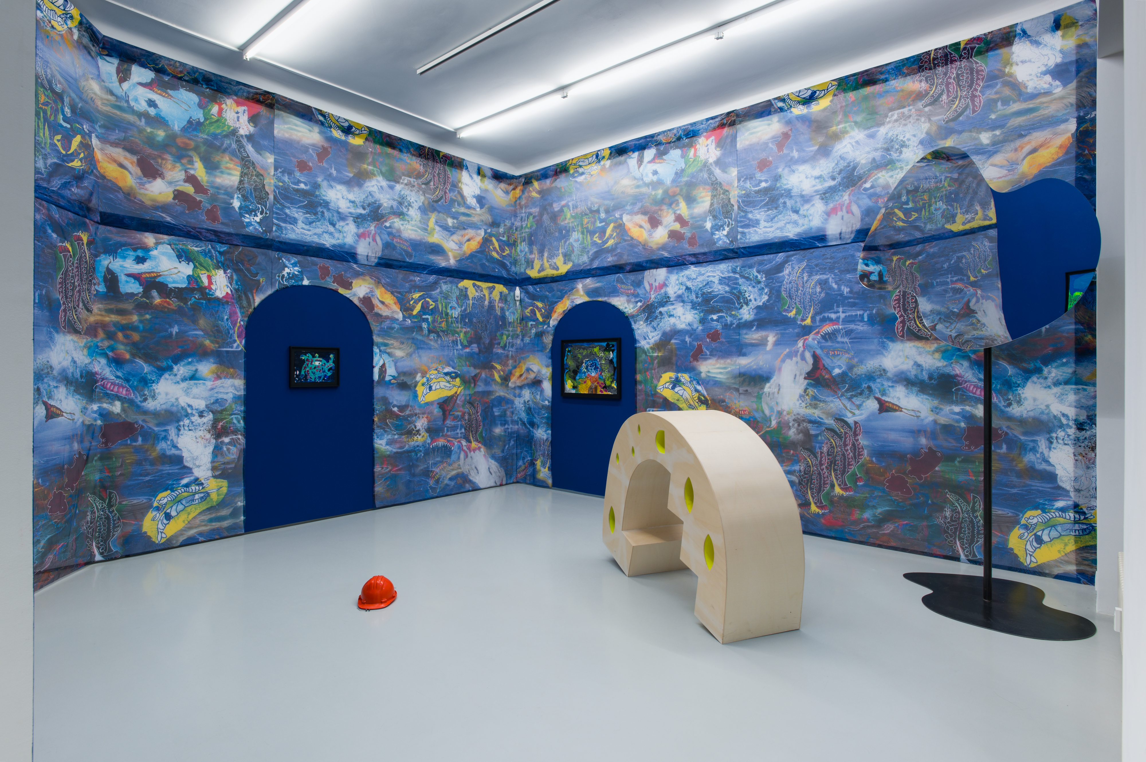 Ein Ausstellungsraum mit bunt gestalteten, großformatig bemalten Wänden, in denen zwei blaue Durchgänge eingelassen sind; im Raum stehen skulpturale Elemente, darunter eine helle, bogenförmige Sitzstruktur und ein roter Schutzhelm auf dem Boden.