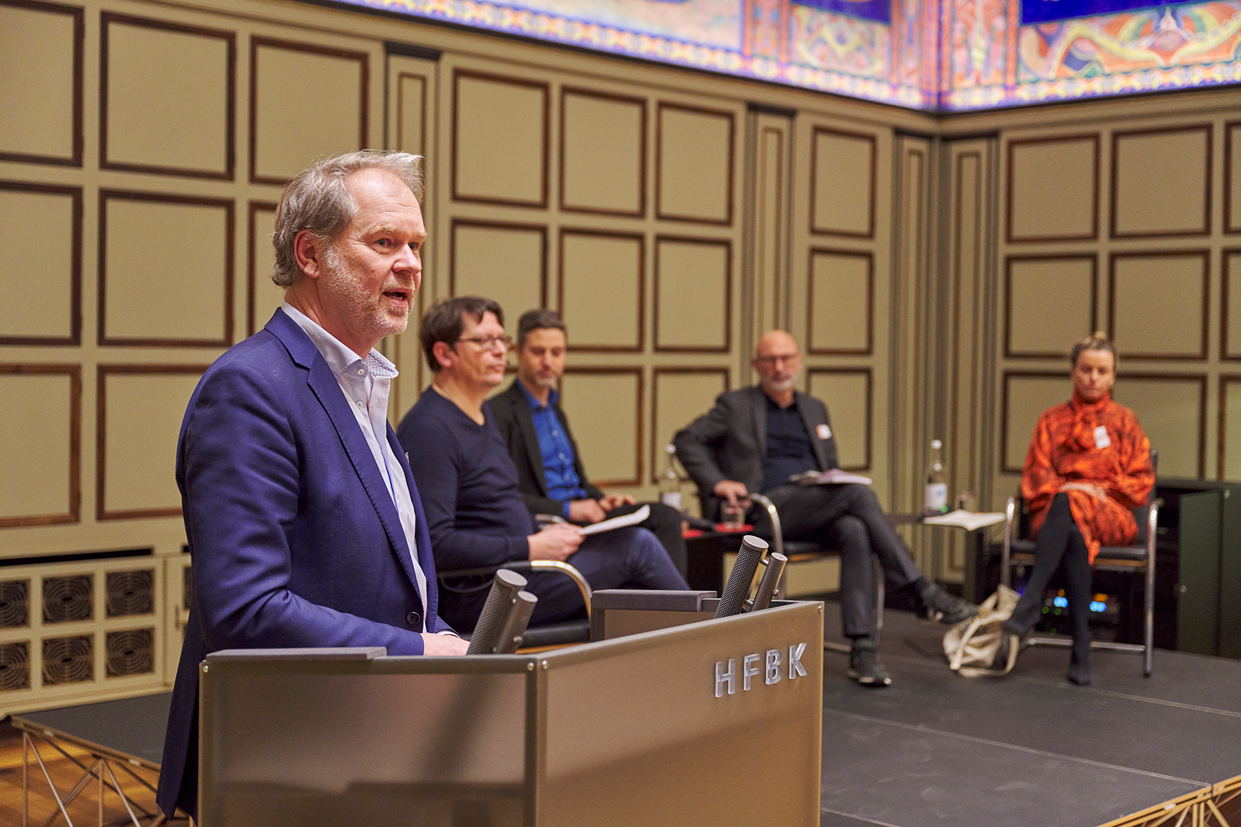 Panel discussion in the auditorium of HFBK Hamburg: a speaker addresses the audience at a lectern while several panelists sit on stage in the background.