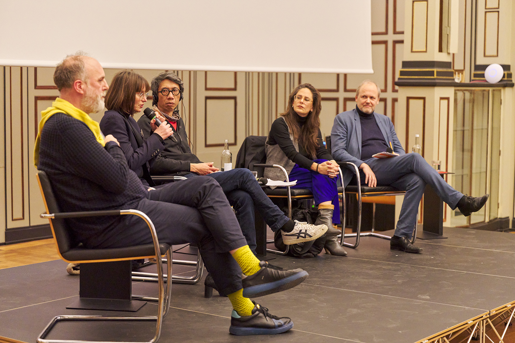 Panel discussion in the auditorium of HFBK Hamburg: five people are seated on stage, one speaking into a handheld microphone while the others listen attentively.