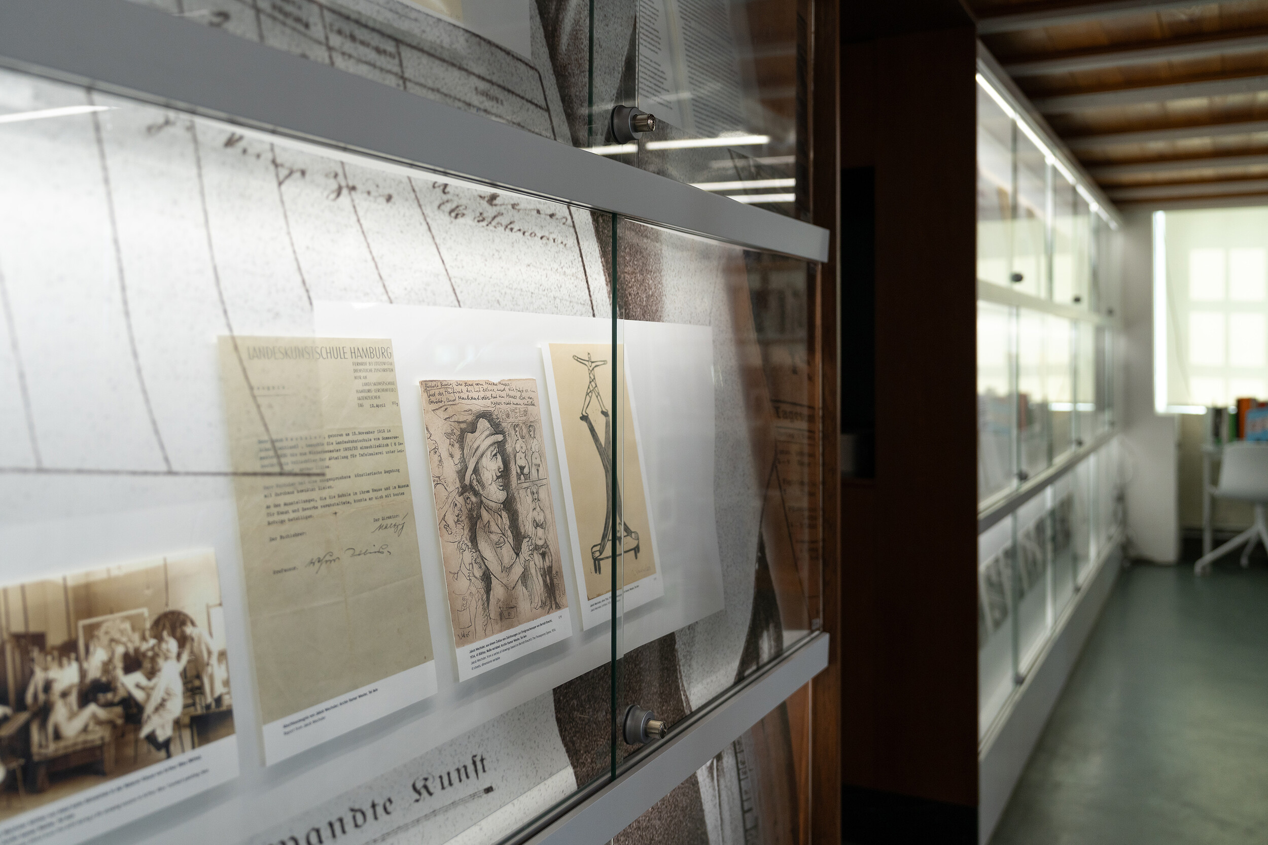 Blick in eine Vitrine der Bibliothek mit historischen Archivdokumenten und Fotografien zur Geschichte der Hochschule für bildende Künste Hamburg im Nationalsozialismus. Die Dokumente sind hinter Glas präsentiert und Teil einer Ausstellung im Bibliotheksmagazin