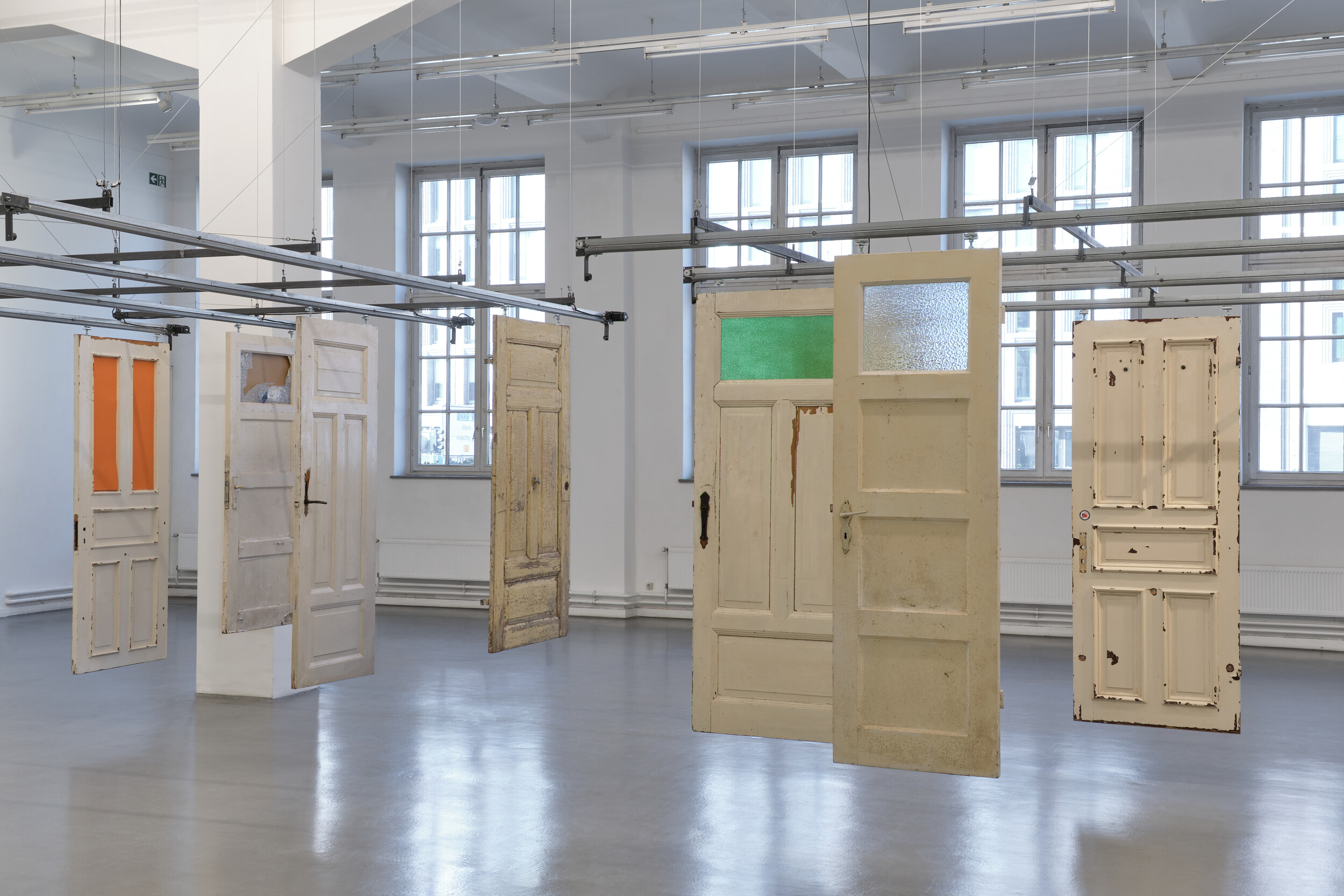 In a bright exhibition space with large windows, several old, differently styled doors hang freely in the room from a metal structure. Some doors feature colored or textured glass panels, while others show signs of wear and peeling paint.