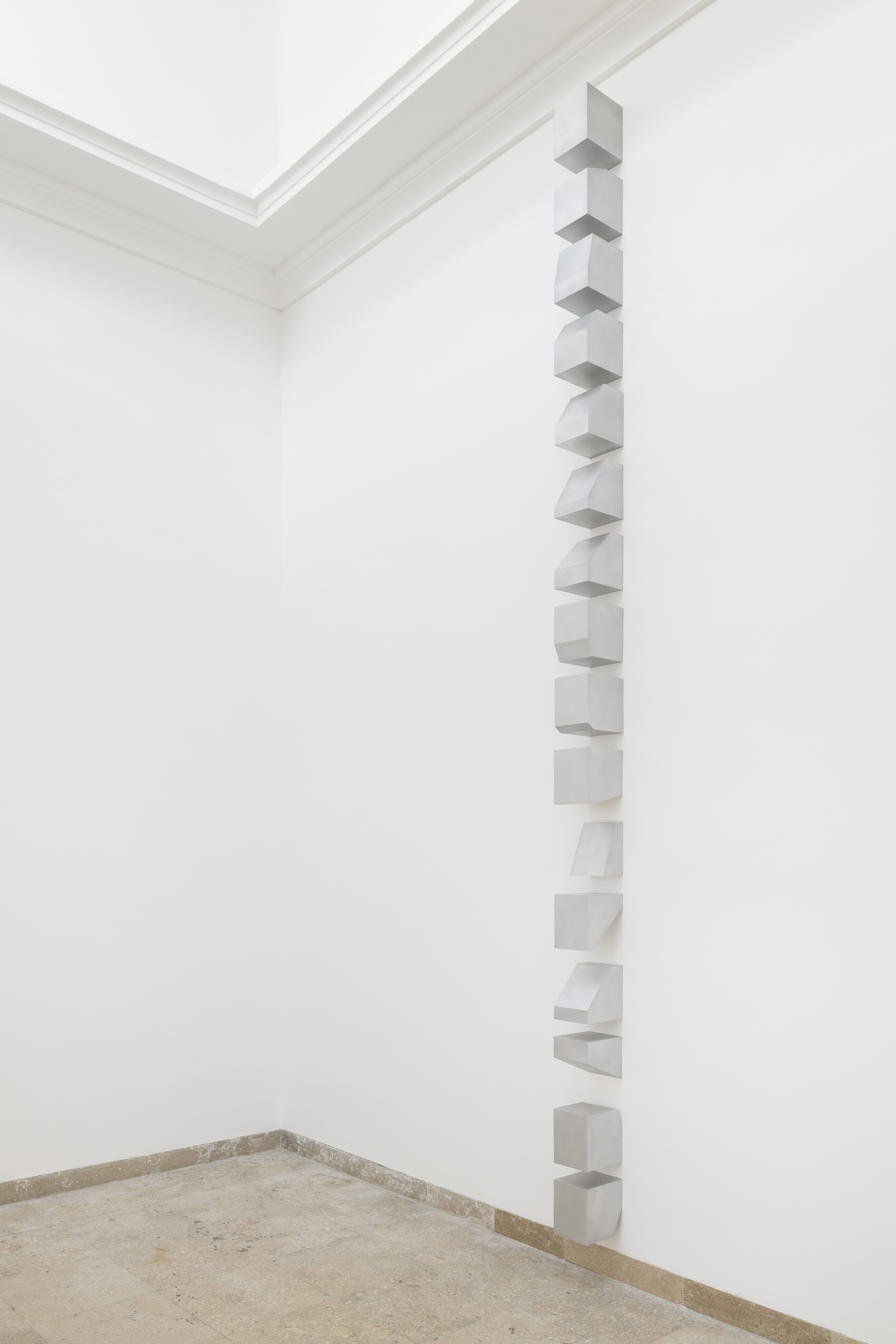 Eine vertikale Reihe weißer, geometrischer Körper verläuft vom Boden bis nahe zur Decke entlang einer weißen Wand. Die einzelnen Elemente ähneln Würfeln oder Quadern, sind jedoch leicht gekippt, verzerrt oder geöffnet und erzeugen einen Rhythmus aus Wiederholung und Variation. Die minimalistische Installation hebt die Beziehung zwischen Form, Raum und Architektur hervor.
