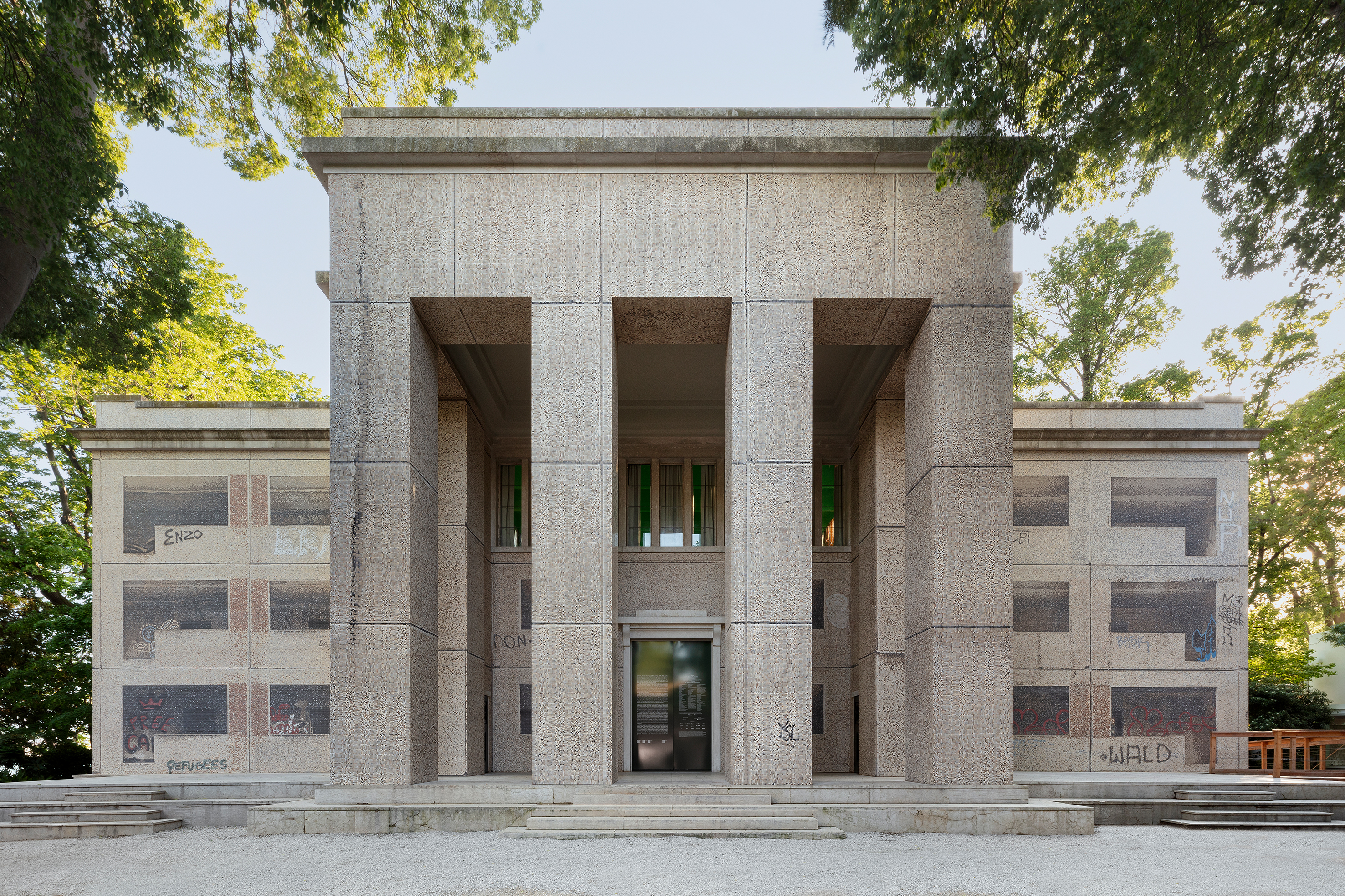 Frontalansicht eines monumentalen Gebäudes mit einer streng symmetrischen Fassade aus hellem Naturstein. Vier massive Pfeiler rahmen den zentralen Eingang, während die Seitenwände mit großflächigen fotografischen Bannern verhängt sind, die die Architektur eines Plattenbaus zeigen und von Graffiti überlagert werden. Das Gebäude ist von hohen Bäumen umgeben und liegt an einem hellen Kiesplatz. Die Intervention verbindet historische Architektur mit zeitgenössischen Spuren urbaner Aneignung.