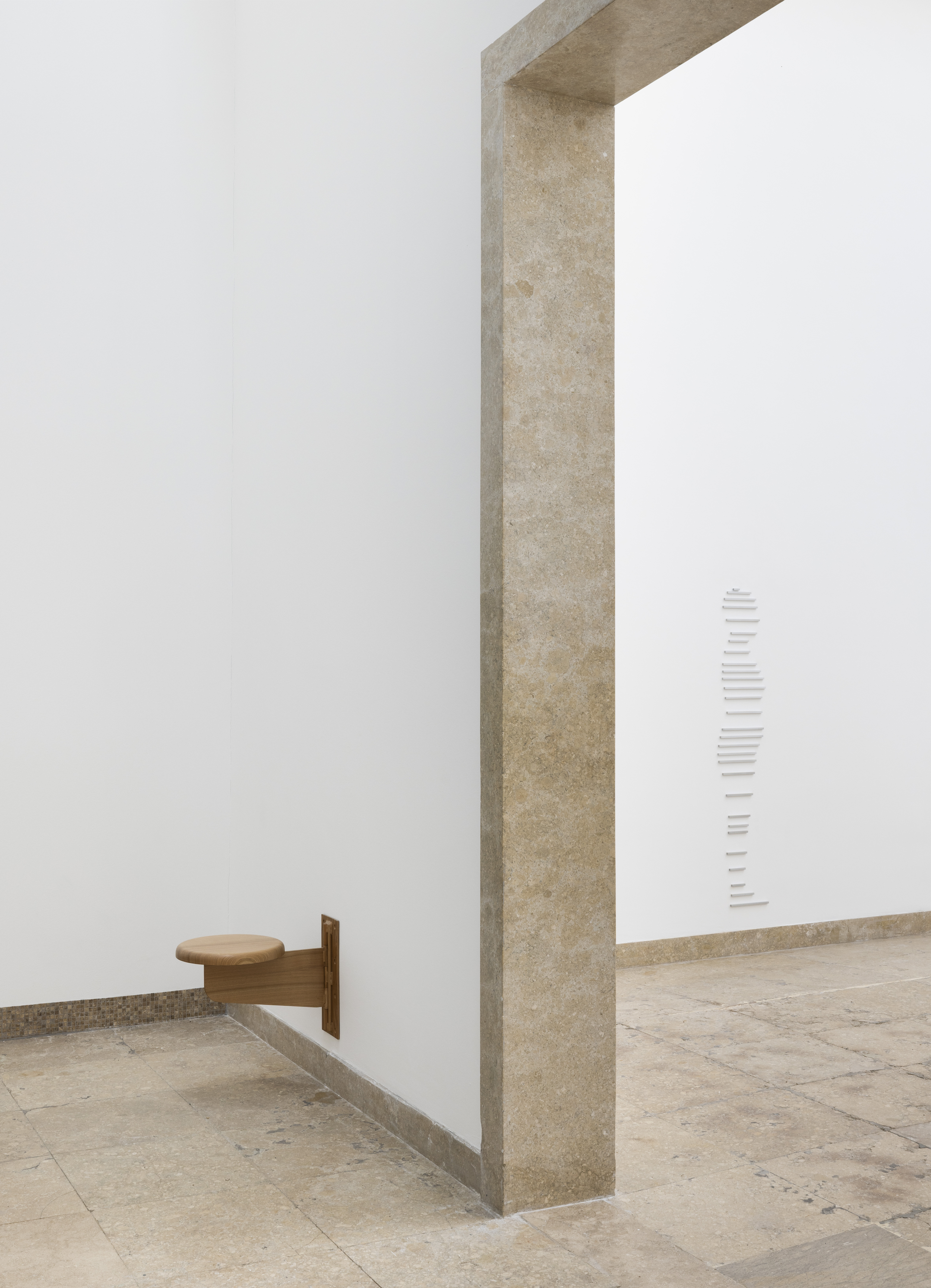 Ein kleiner runder Holzsitz ragt aus einer weißen Wand in einen nahezu leeren Ausstellungsraum hinein. Ein breiter Steinrahmen markiert den Übergang zu einem angrenzenden Raum, in dem eine vertikale Anordnung schmaler weißer Elemente an der Wand zu sehen ist. Die minimalistische Installation verbindet skulpturale Interventionen mit architektonischen Strukturen und lenkt die Aufmerksamkeit auf Raum, Proportion und Wahrnehmung.