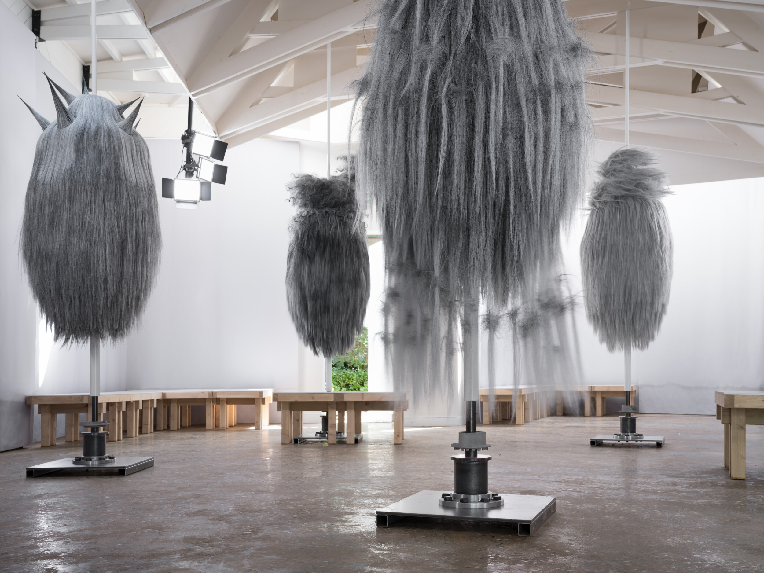 Exhibition view featuring four larger-than-life, densely fur-covered figures mounted on metal stands in a bright space; the figure on the left has several pointed horns.