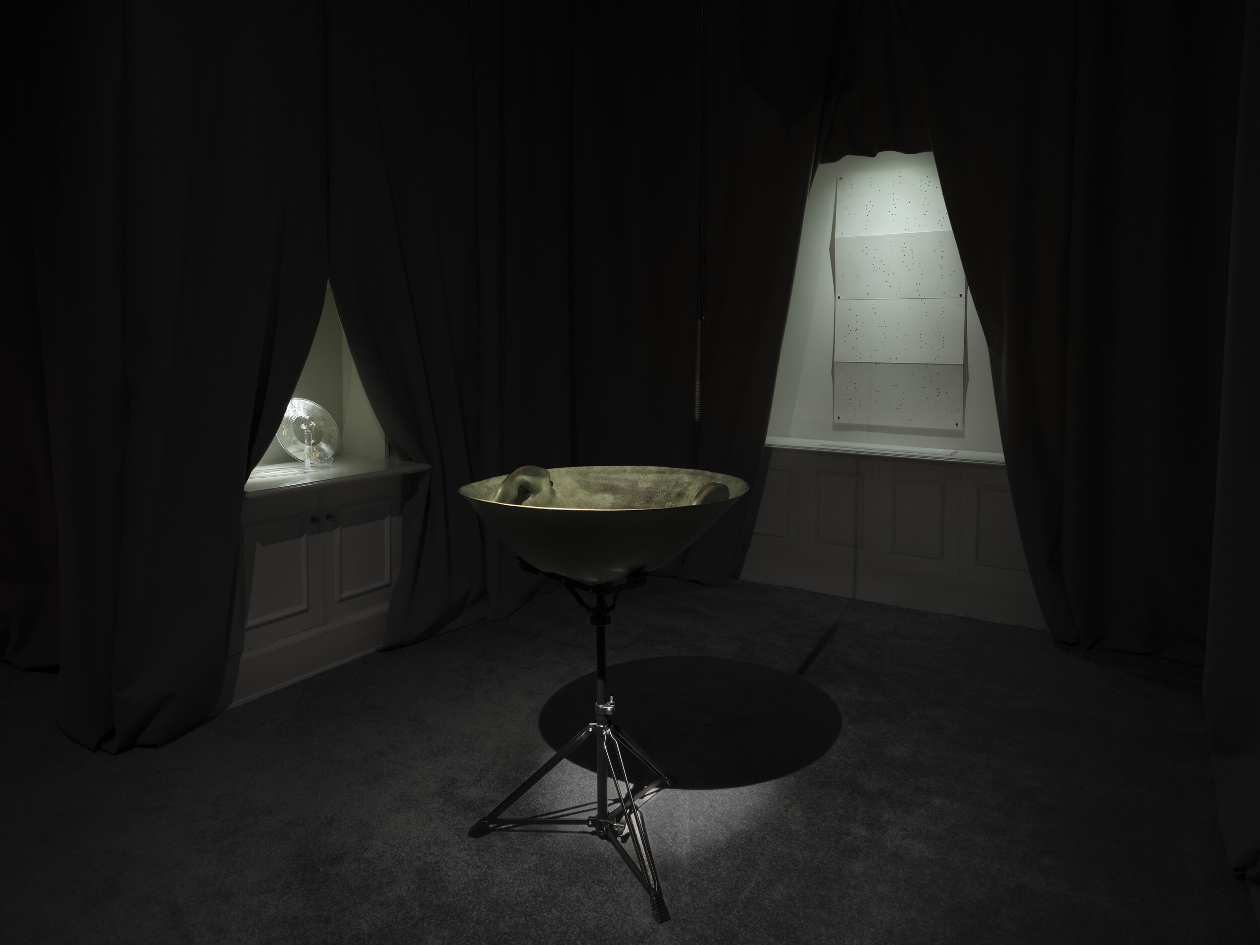Dark room installation with black curtains and a bowl-shaped sculpture mounted on a metal stand. A fan and a sheet mounted on the wall are visible through openings in the curtains.