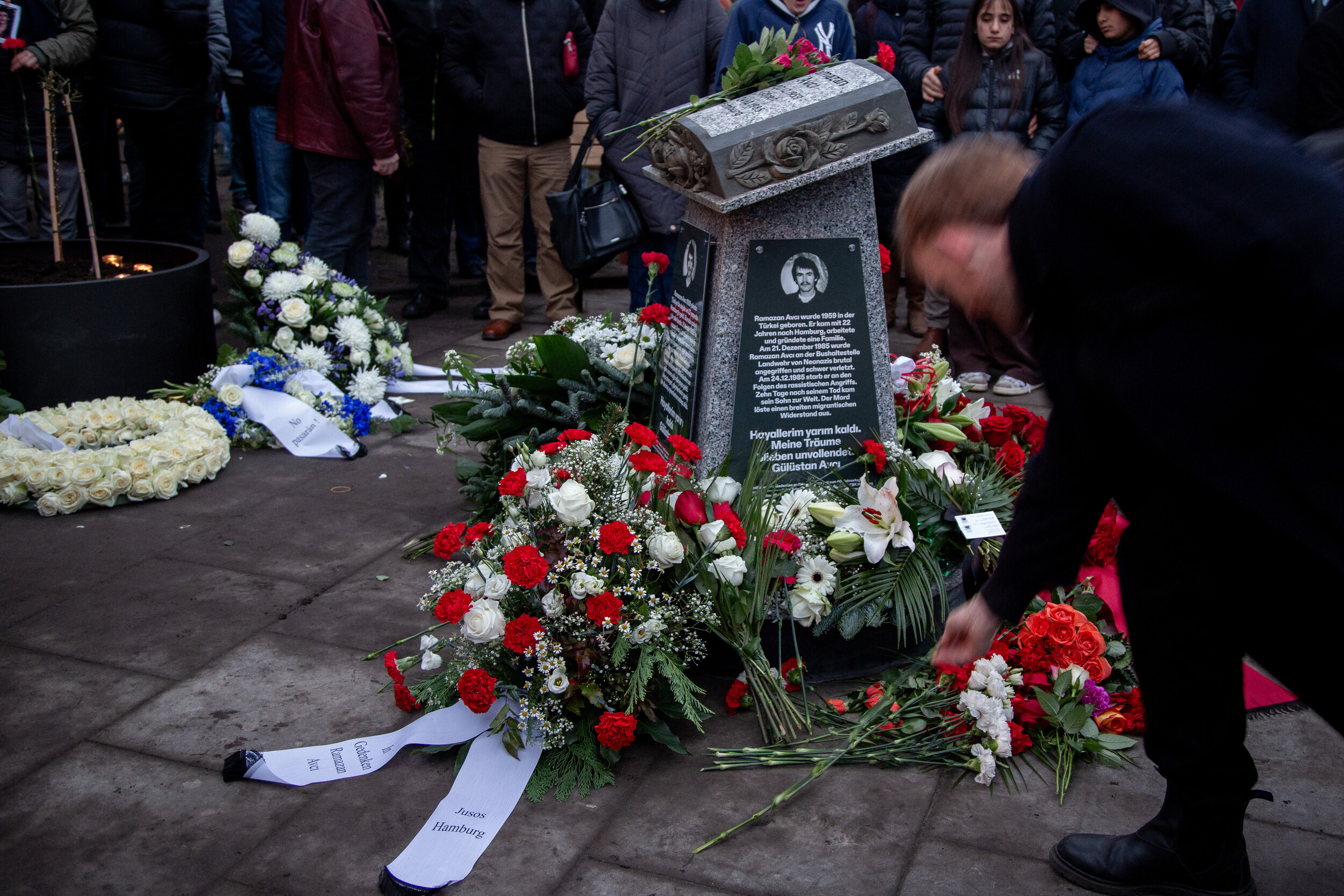 Gedenkveranstaltung an einem Denkmal für Ramazan Avcı, umgeben von zahlreichen Blumen und Kränzen. Menschen stehen im Hintergrund, während im Vordergrund weitere Blumen niedergelegt werden.