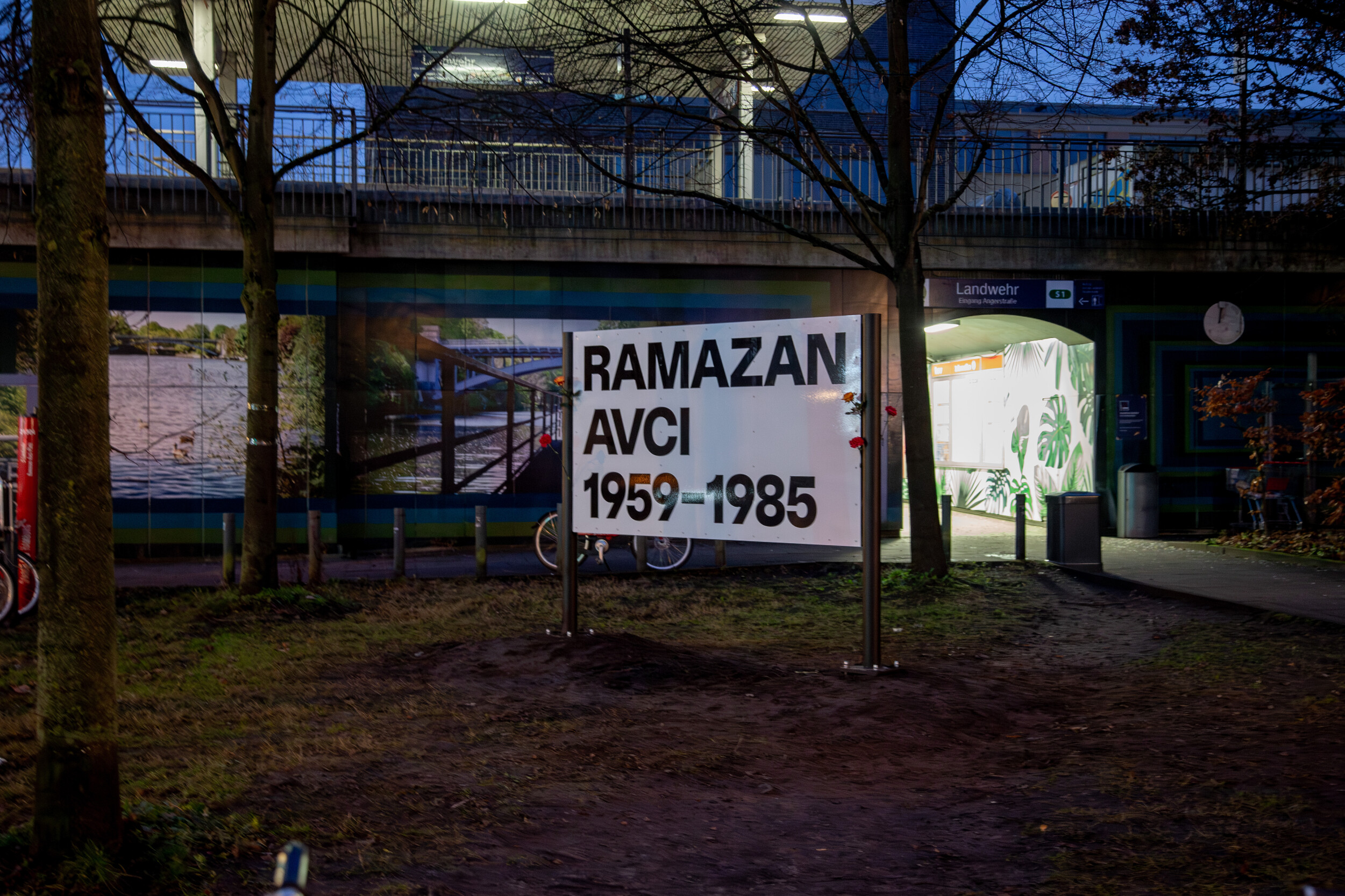 Auf einem Vorplatz vor einem S-Bahn-Eingang ist ein Schild auf Stehlen zu sehen mit der Aufschrift "Ramazan Avcı 1959-1985".