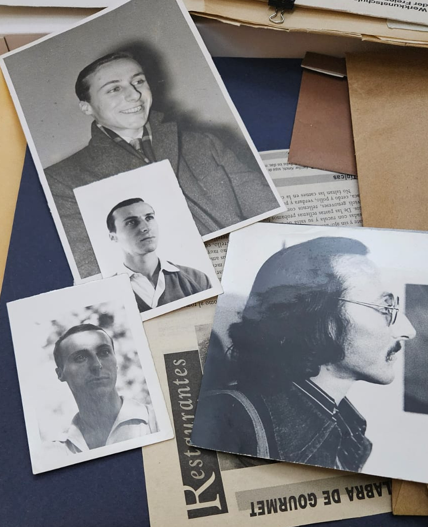 Several black-and-white portrait photographs of a man are arranged on a table, surrounded by papers and envelopes. The images show him in different poses and perspectives.