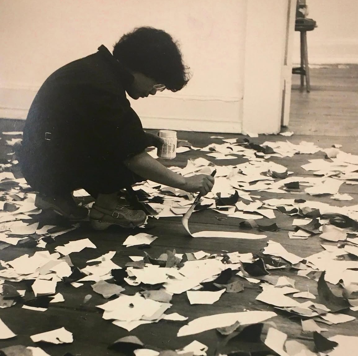 Black-and-white photograph of a person kneeling on the floor, working with a brush or tool on a dark surface. The floor is covered with torn paper pieces; an empty interior space is visible in the background.
