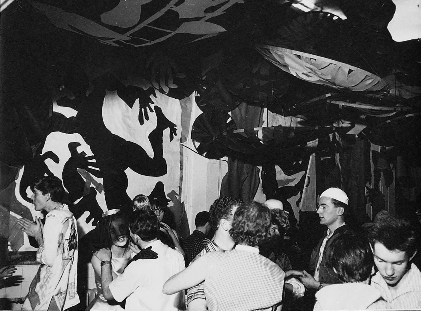 Black-and-white photograph of a crowded group of people in an interior space, likely at a party or event. Large, high-contrast shadow or cut-out figures cover the walls and ceiling, creating a dynamic, almost surreal atmosphere.