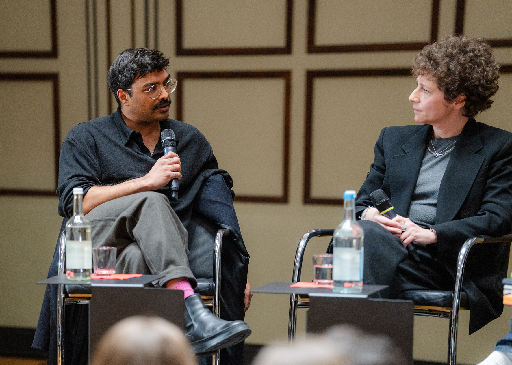 Two people are seated on a stage, engaged in conversation. Both are holding handheld microphones and facing each other. Small tables with water bottles and glasses are placed in front of them. A geometrically patterned wall is visible in the background, with the audience blurred in the foreground.