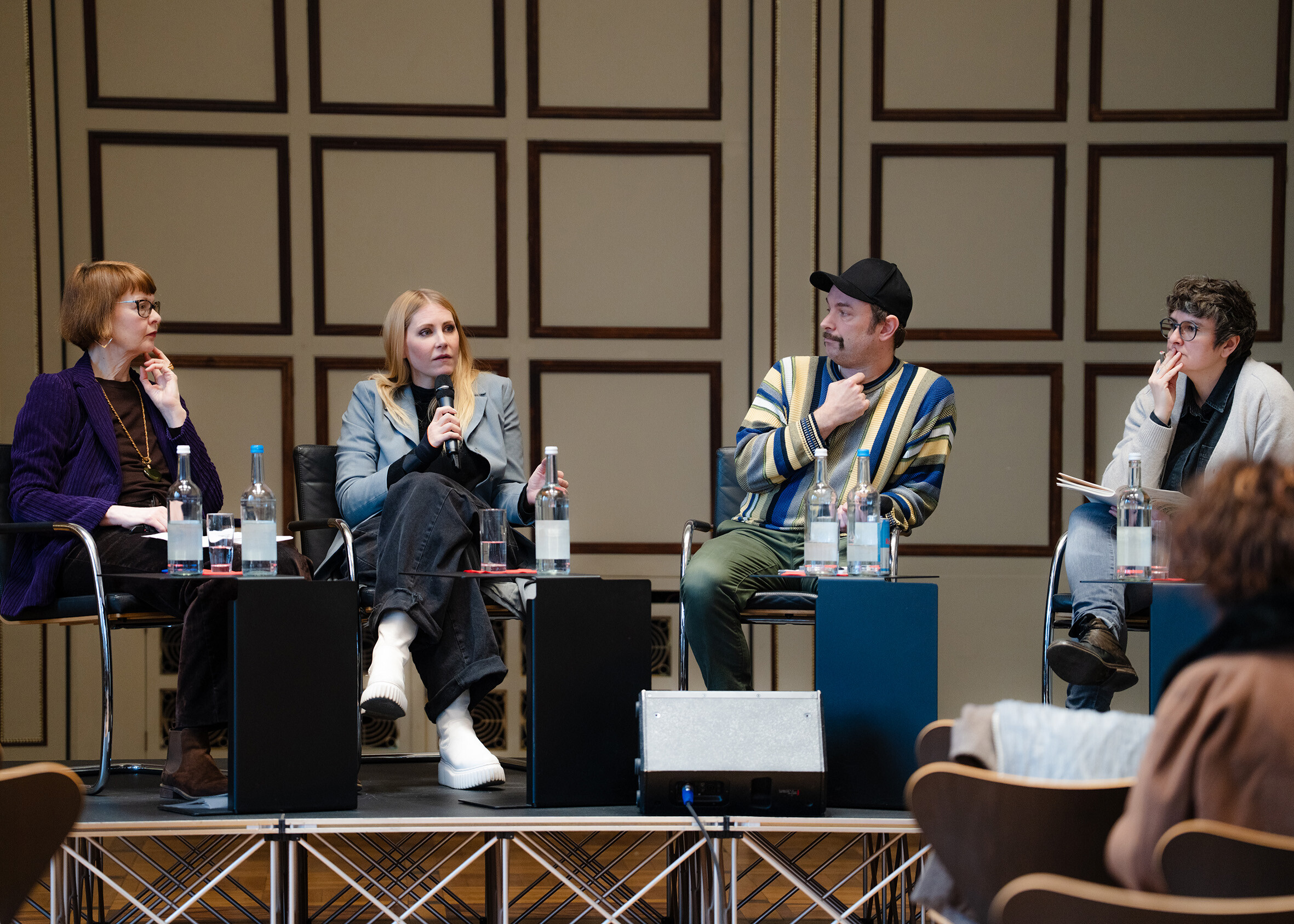 Four people are seated on a stage participating in a panel discussion. One person is speaking into a handheld microphone while the others listen or take notes. Small tables with water bottles are placed in front of them. Parts of the audience are visible in the foreground, with a geometrically patterned wall in the background.