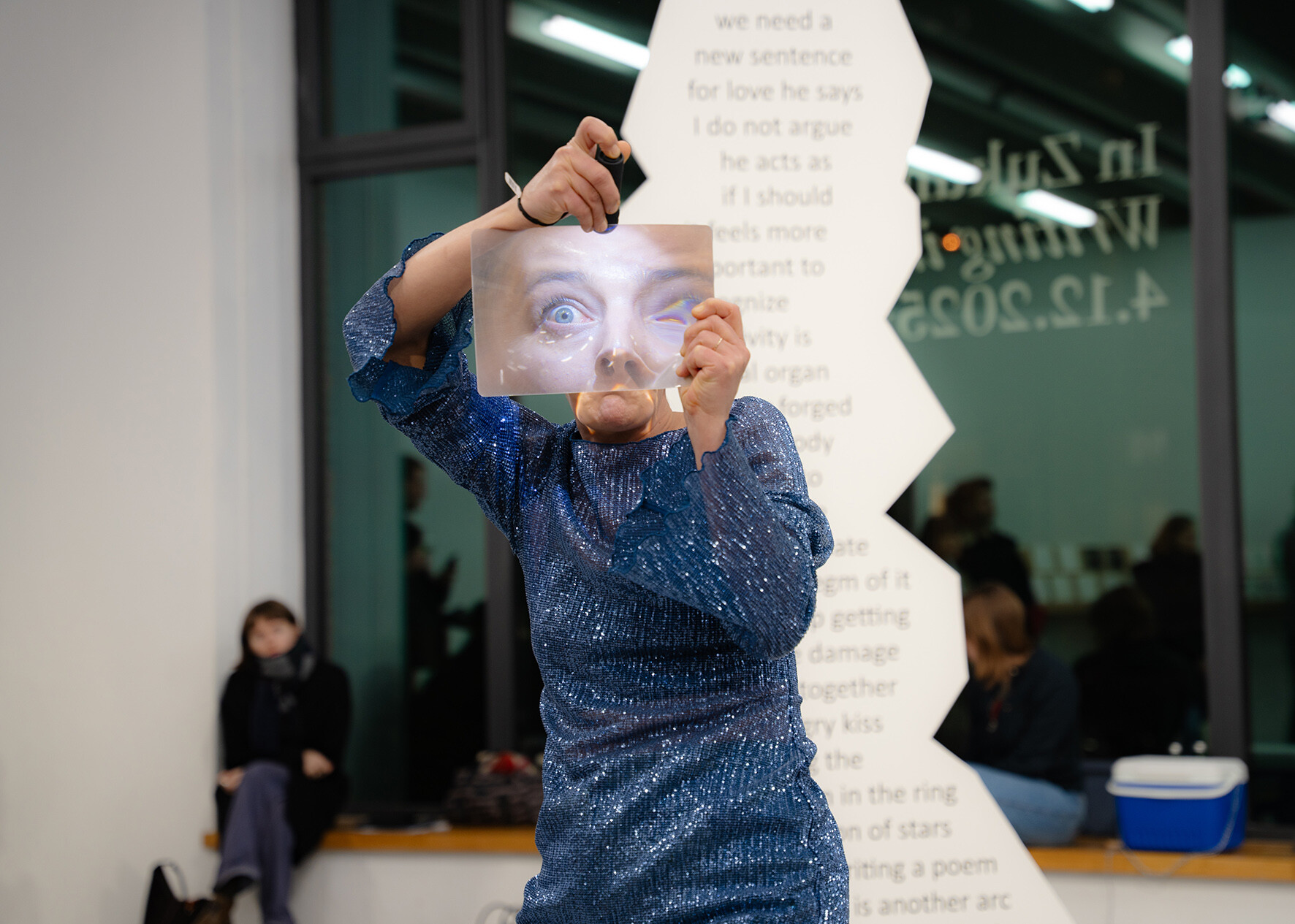 Eine Person in einem glitzernden blauen Oberteil hält eine transparente Folie mit einem vergrößerten Auge vor ihr Gesicht; im Hintergrund sitzen Menschen in einem Innenraum mit Fenstern und Textinstallation.
