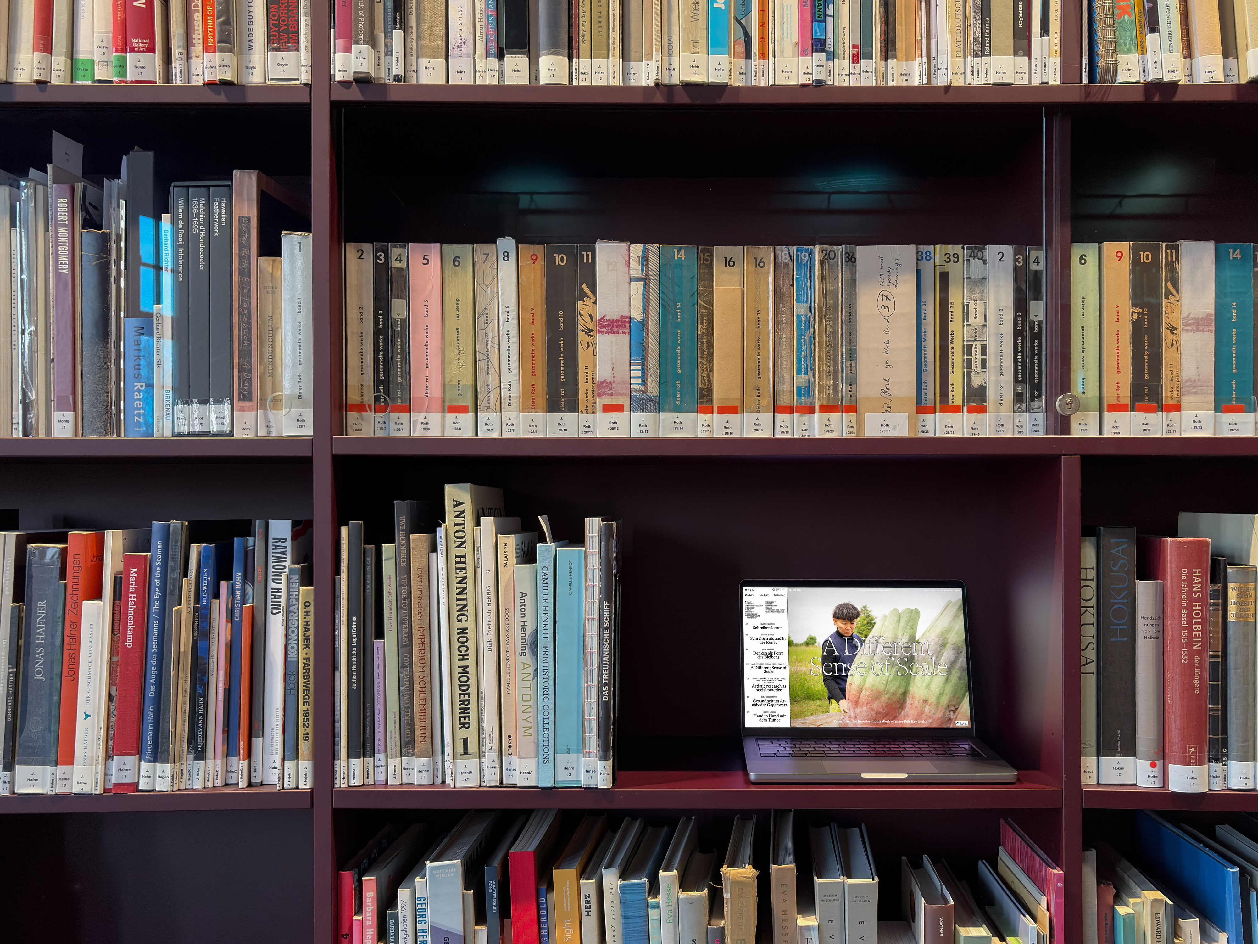 A library bookshelf filled with tightly arranged books across several shelves; on one shelf, an open laptop displays a person outdoors on its screen.