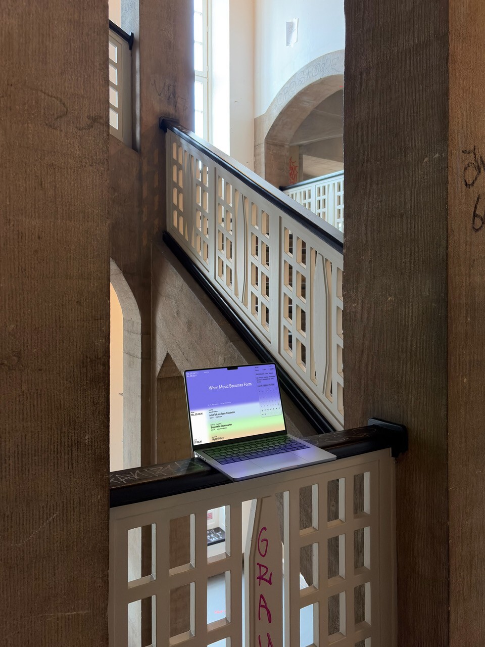 Ein offener Laptop steht auf einem Treppengeländer in einem hellen Innenraum mit hohen Fenstern, Holzsäulen und einer dekorativen weißen Brüstung; auf dem Bildschirm ist eine Benutzeroberfläche mit Text zu sehen.