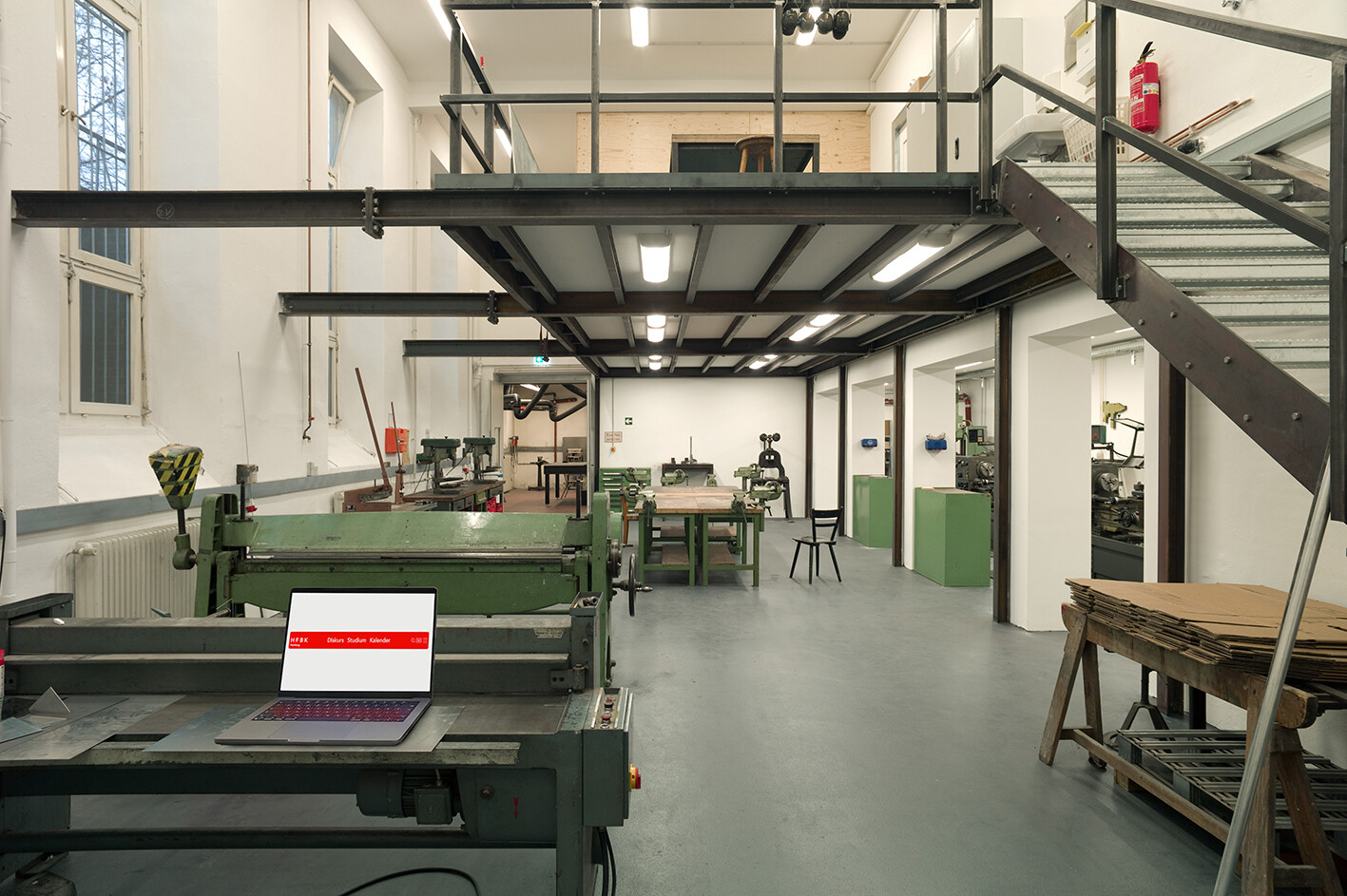 Eine Werkstatt mit industriellen Maschinen, Arbeitstischen und einer Metallempore; im Vordergrund steht ein Laptop auf einer großen grünen Maschine, während im Raum verschiedene Geräte und Materialien verteilt sind.