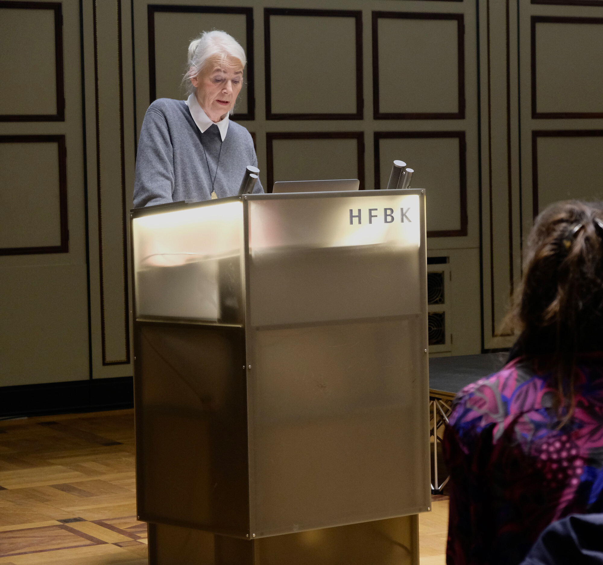 A woman stands at a lectern and reads a text to an audience.