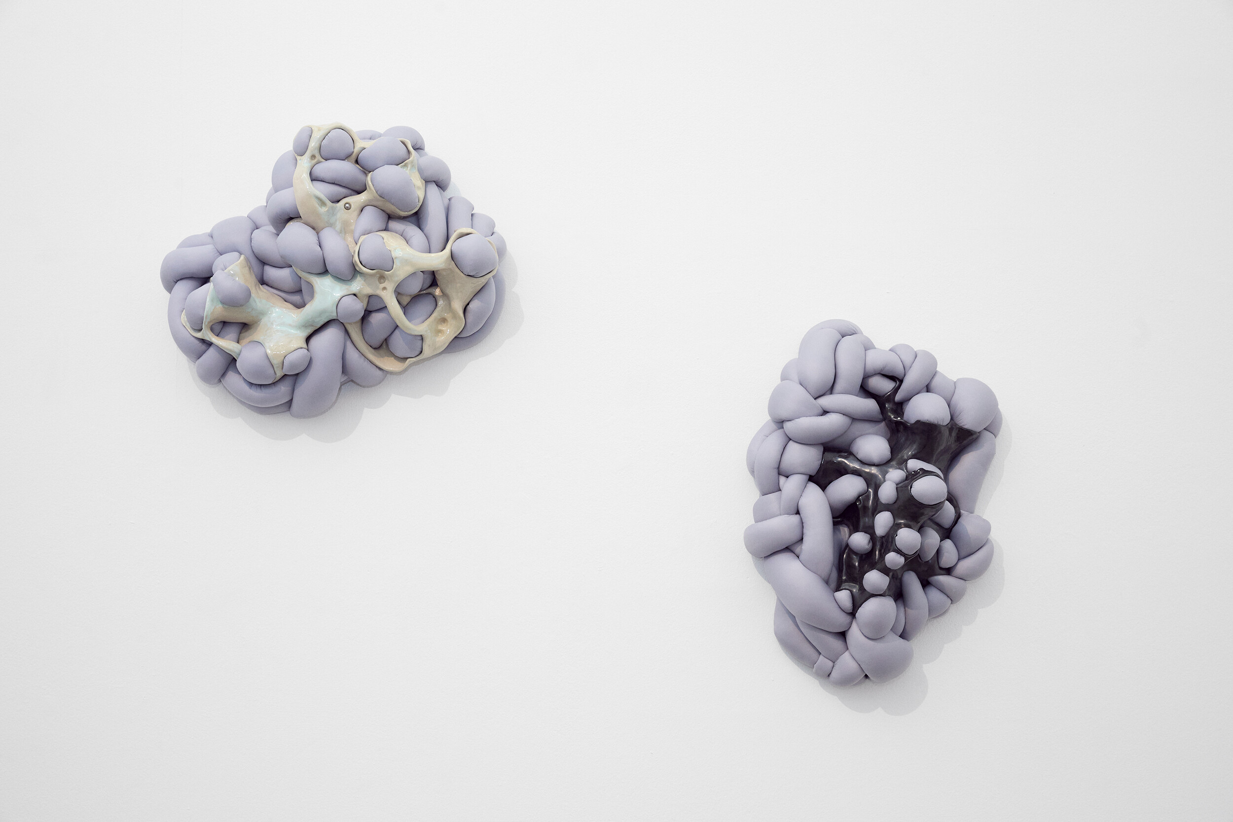 Zwei abstrakte, organisch geformte Wandobjekte in hellvioletten Tönen, die aus verschlungenen, knotenartigen Formen bestehen und auf einer weißen Wand angebracht sind.