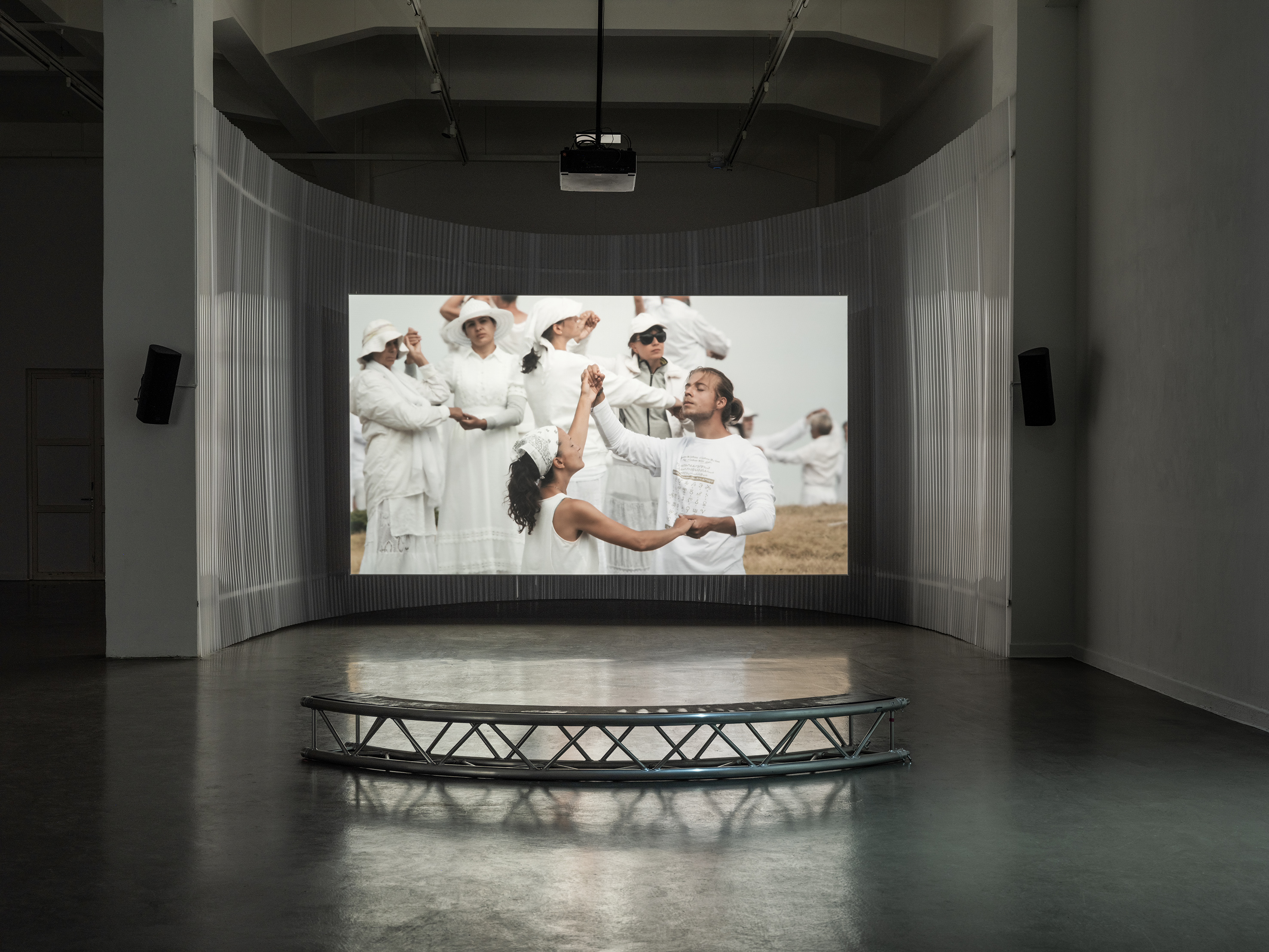 Ausstellungsansicht einer Videoinstallation mit gebogener Projektionsfläche und einer niedrigen, halbkreisförmigen Sitzbank. Das projizierte Bild zeigt eine Gruppe weiß gekleideter Menschen beim gemeinsamen Tanz.