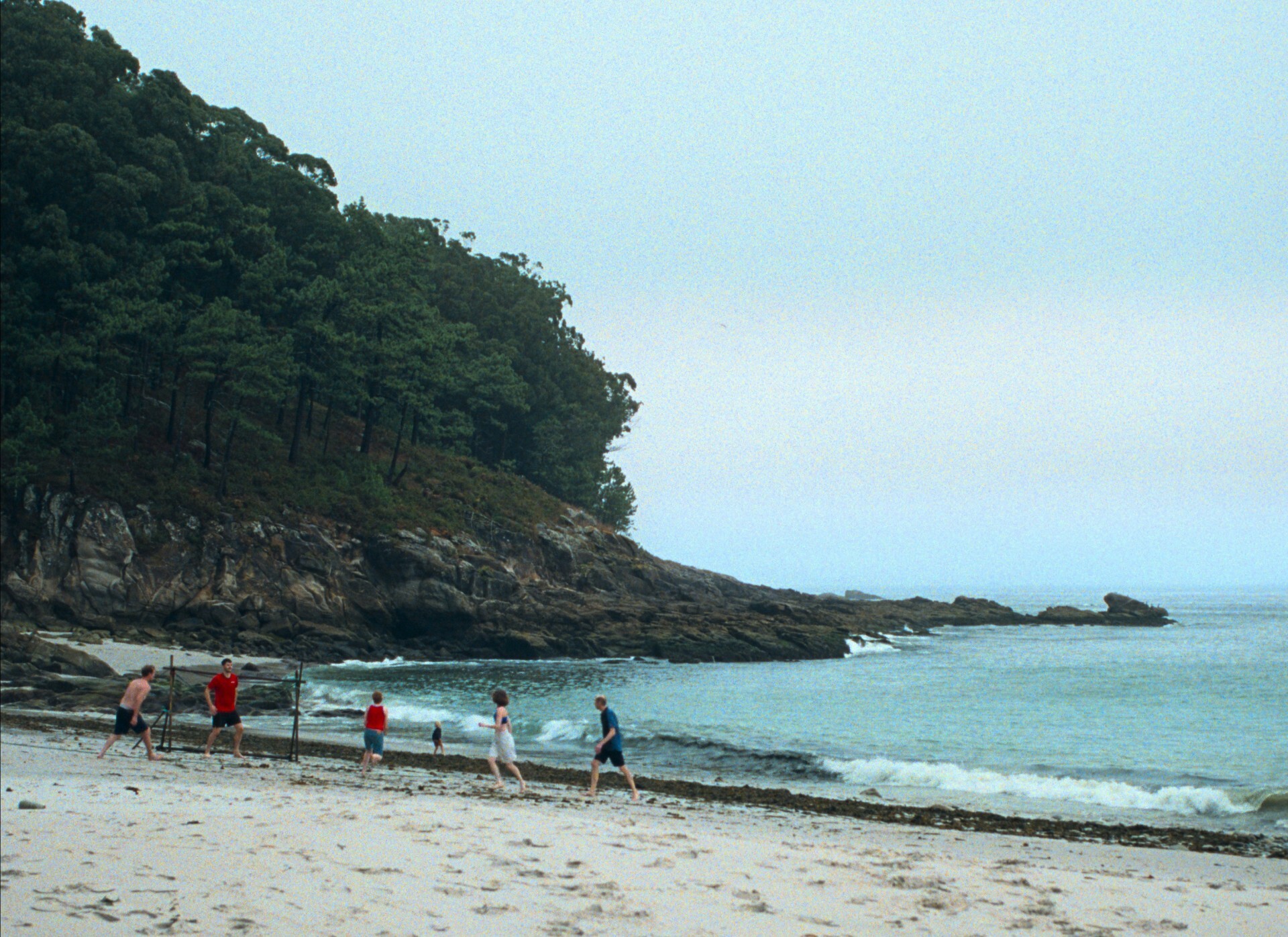 Mehrere Menschen spielen an einem bewölkten Tag Fußball an einem ruhigen Sandstrand. Im Hintergrund ragen bewaldete Felsen und eine felsige Küstenlinie ins Meer. Die körnige, leicht entsättigte Bildästhetik erzeugt eine melancholische, filmische Stimmung.