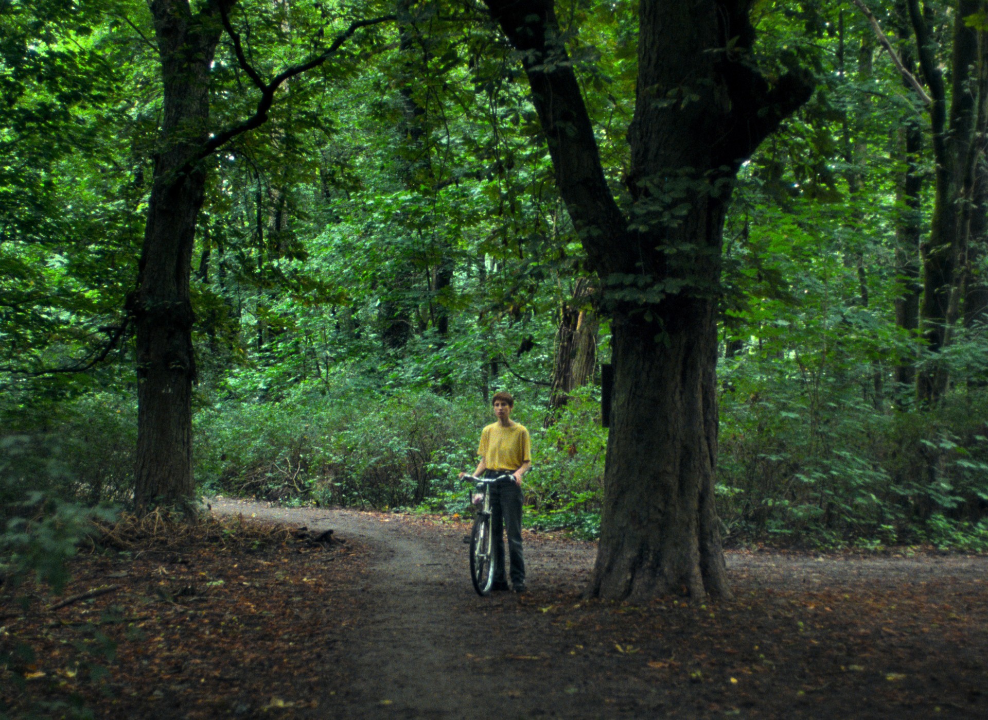 Eine Person steht mit einem Fahrrad auf einem schmalen Waldweg zwischen hohen Bäumen und dichter grüner Vegetation. Das diffuse Licht und die körnige Bildästhetik erzeugen eine ruhige, leicht melancholische Atmosphäre.