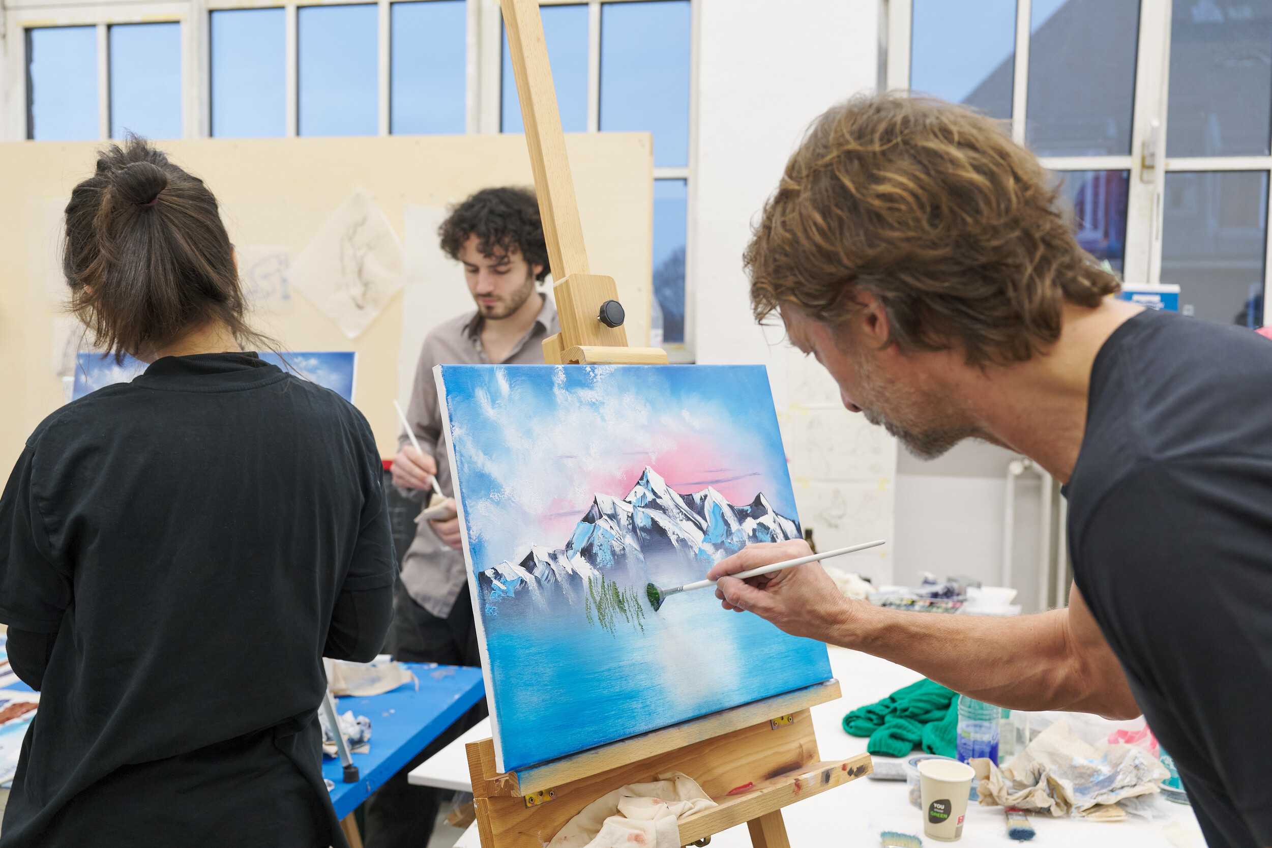 A person paints a landscape with mountains and water on an easel in an art studio, while others work in the background; paints and materials are spread across the tables.