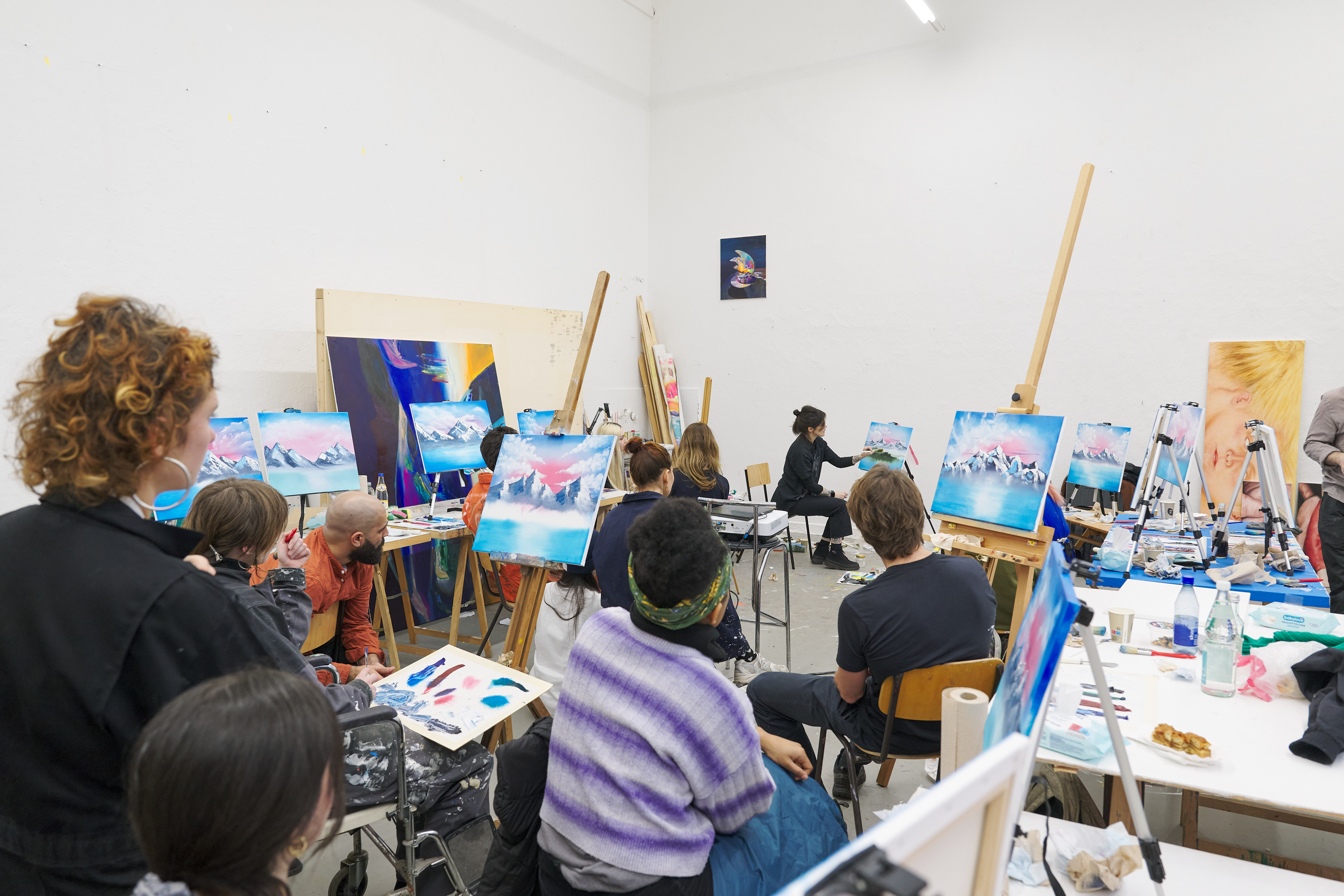 A group of people sits in an art studio watching a painting demonstration; several easels display canvases with landscape scenes in blue and pink tones, while paints and materials are spread across tables.