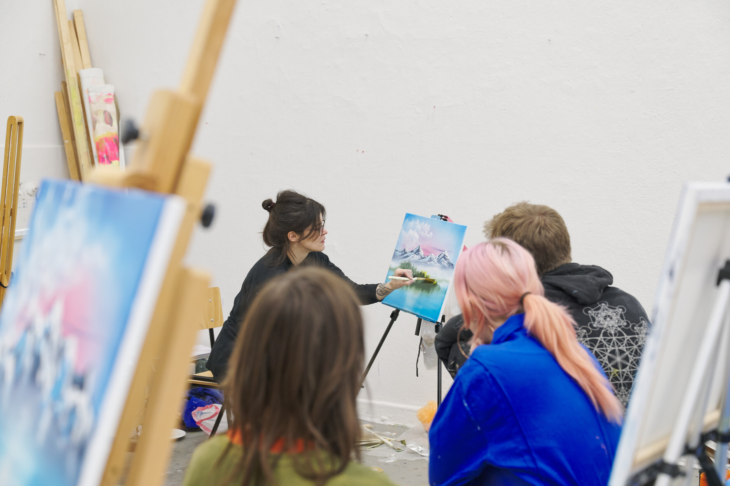 A person demonstrates painting a landscape on a canvas in an art studio, while several participants sit in front watching; easels and additional paintings are arranged around the room.