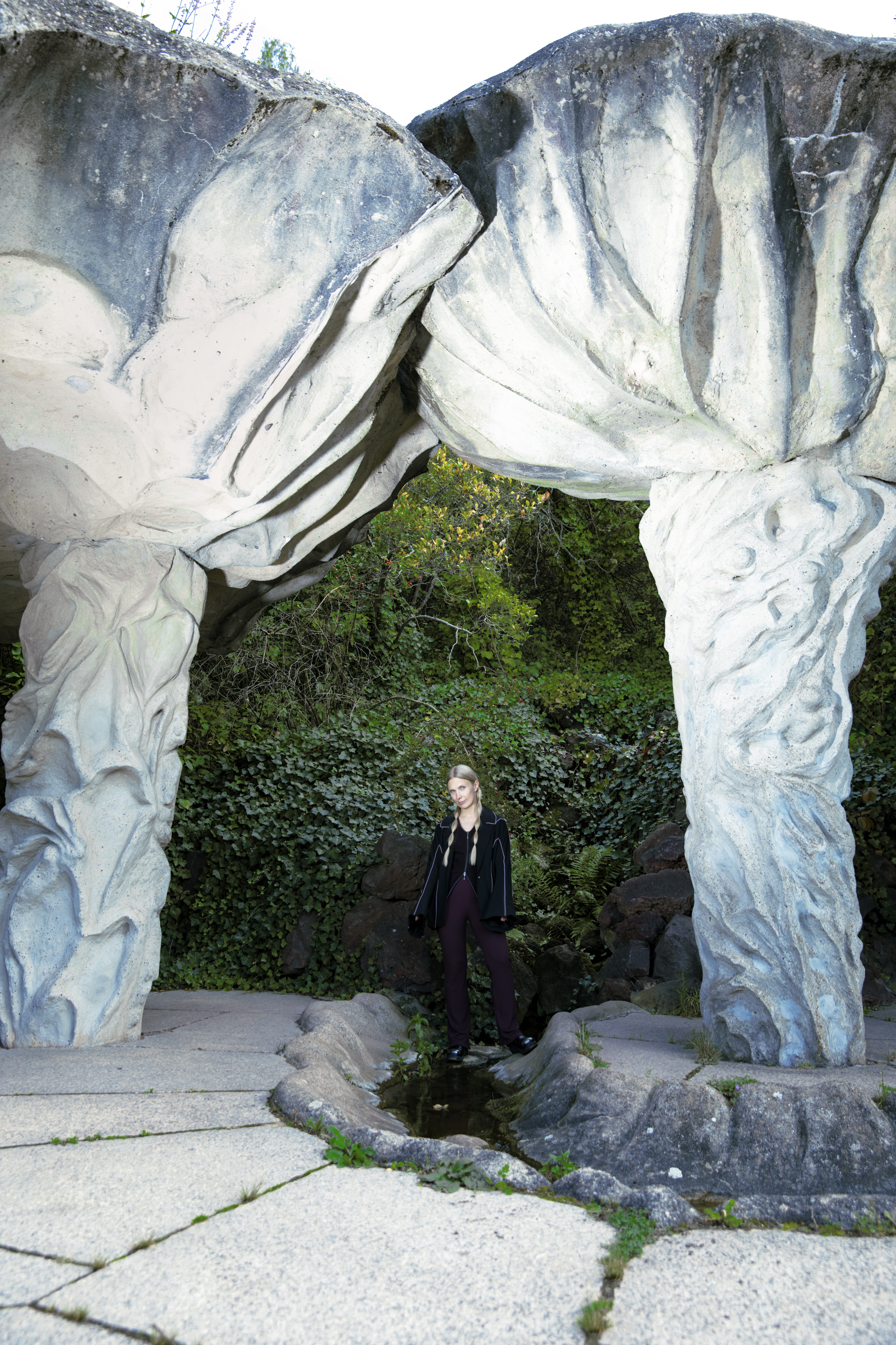 A woman dressed in black with long blonde braids is standing beneath two large, mushroom-shaped stone sculptures in an overgrown outdoor area.