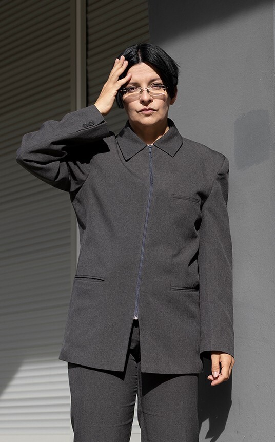 A woman with short dark hair and glasses looks into the camera and holds one hand to her head. She is wearing gray clothing.