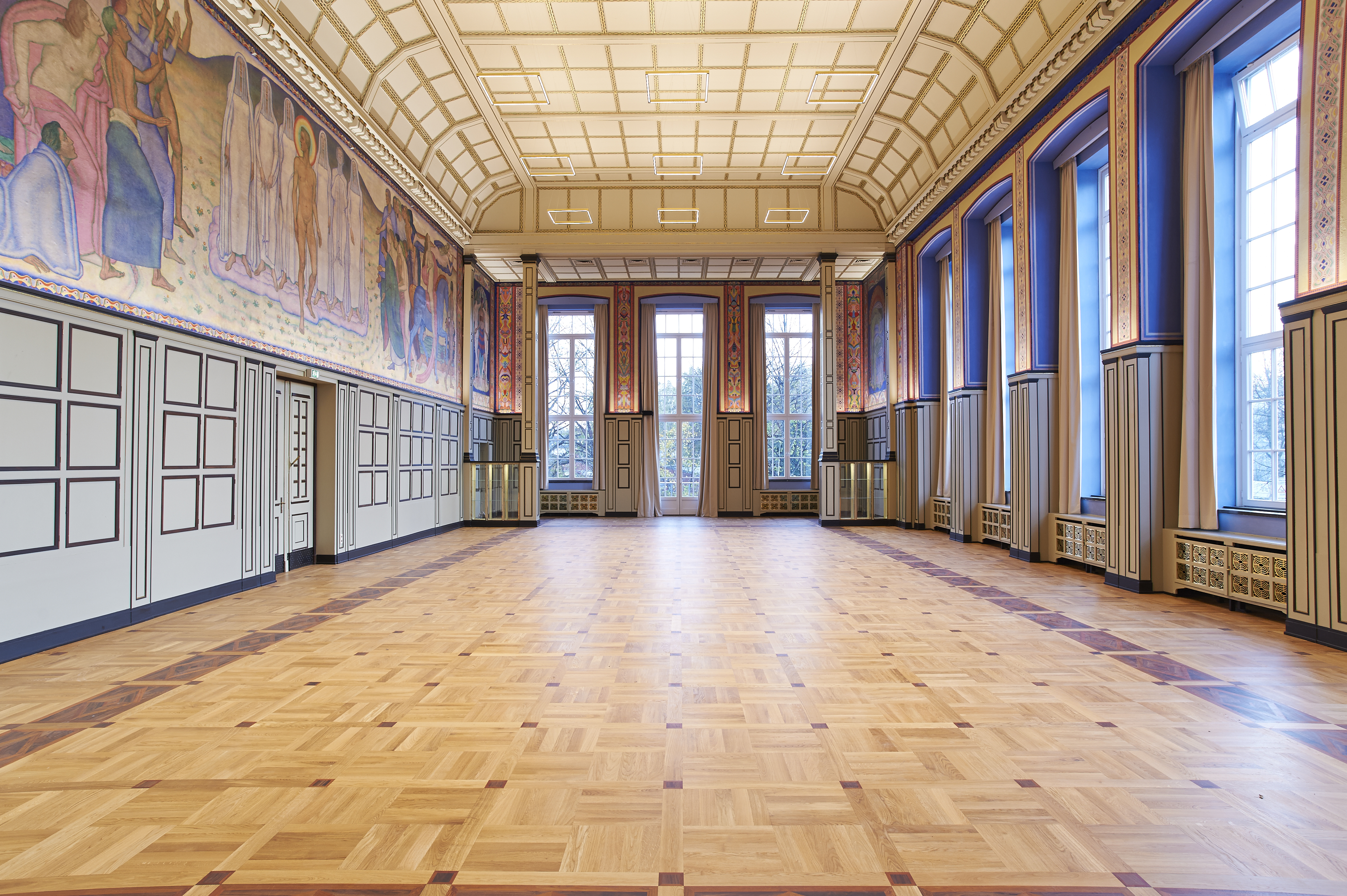 A historic, spacious room with wooden floors, paintings on the walls, large windows, and a coffered ceiling.