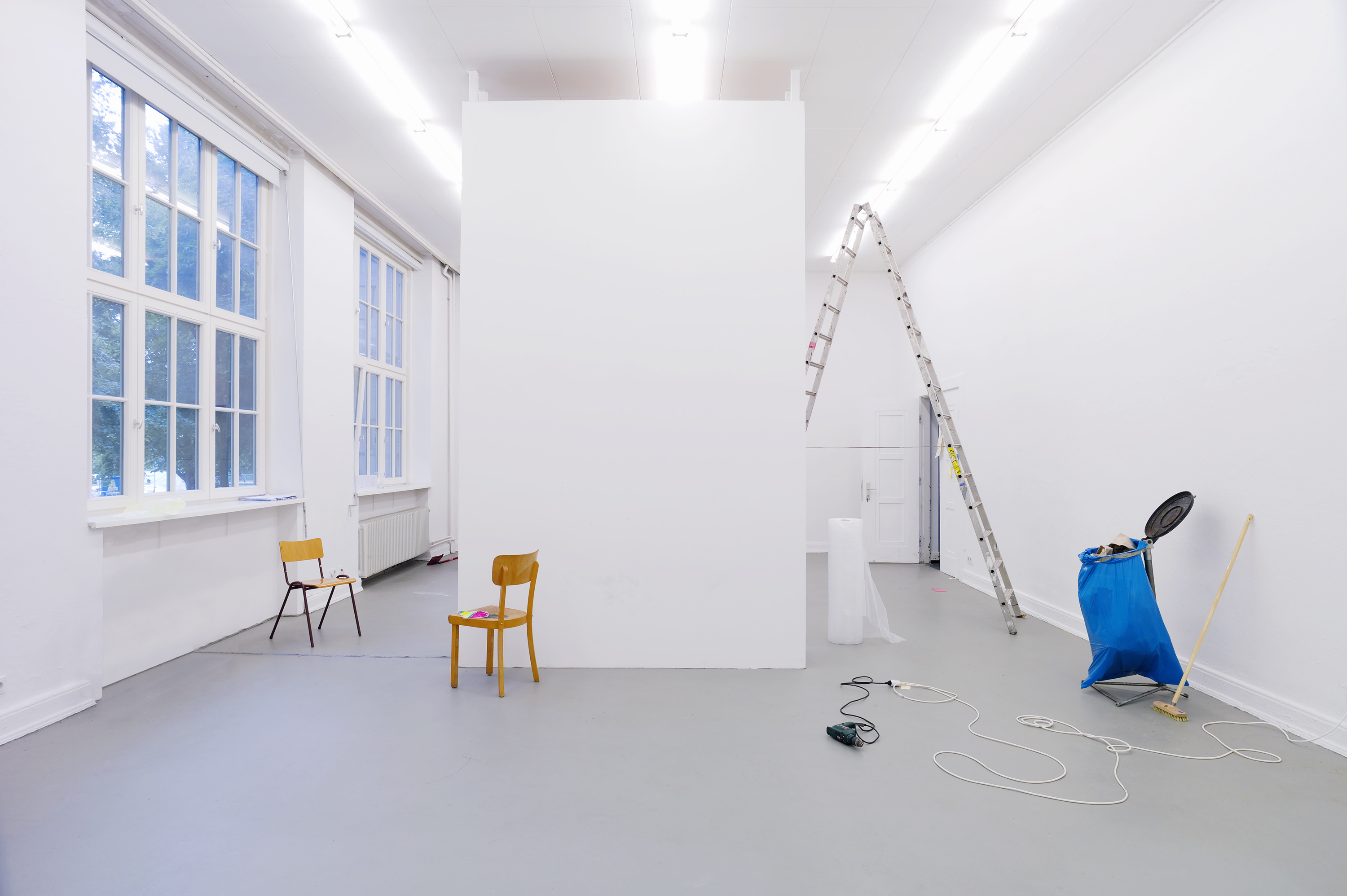 Bright white studio space with high ceilings and large windows on the left. A freestanding white wall stands in the center. Simple wooden chairs are placed in front of and beside it. On the right, a ladder leans against the wall; a cable and a power tool lie on the floor. A blue trash bag, a broom, and a roll of paper suggest installation or setup work.