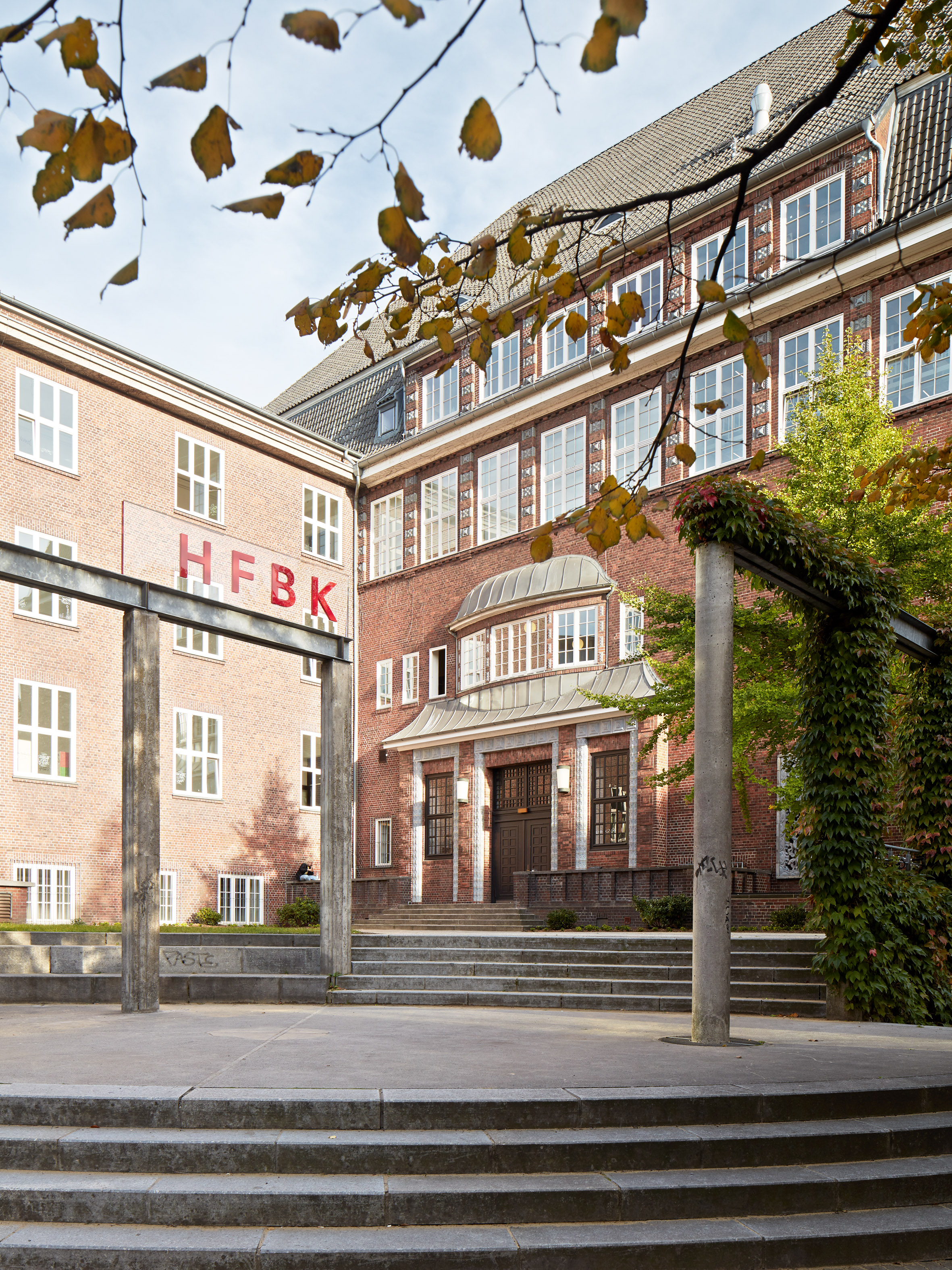 Das Hauptgebäude der HFBK Hamburg aus rotem Backstein mit dem Schriftzug „HFBK“ an der Fassade, gesehen über einen Platz mit Stufen, Säulen und herbstlichen Baumzweigen im Vordergrund.