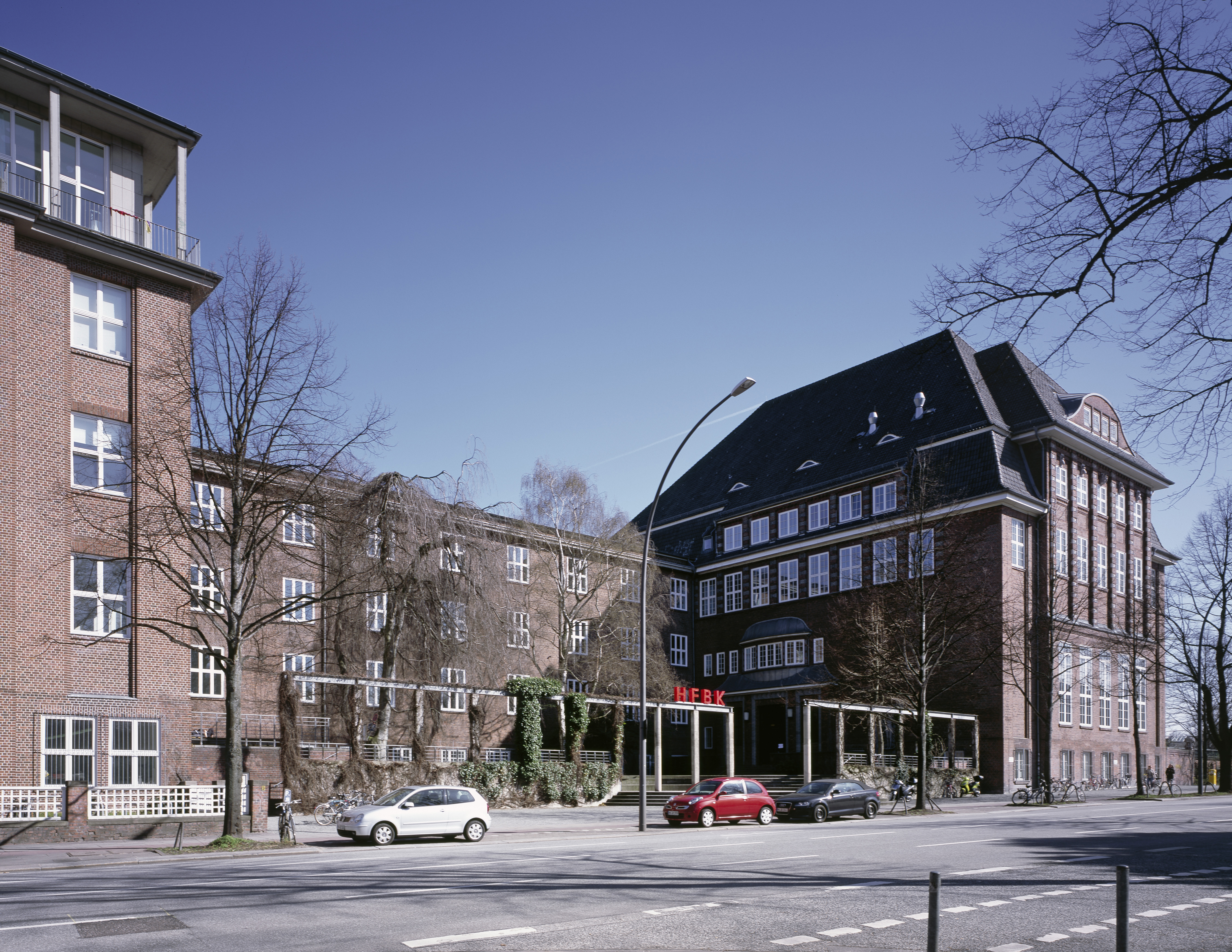 Exterior view of the HFBK Hamburg: a large, historic brick building with several wings, many windows, and a distinctive entrance. In front of it runs a street with parked cars and bicycles; leafless trees frame the building beneath a blue sky.