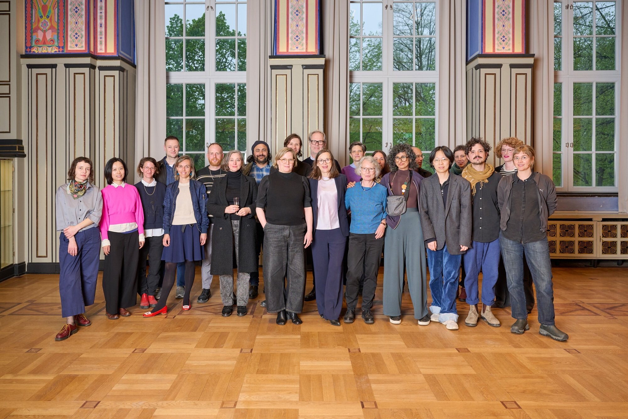 Large group of around twenty people standing side by side in a formal hall with tall windows, decorated columns, and parquet flooring; all are facing the camera.