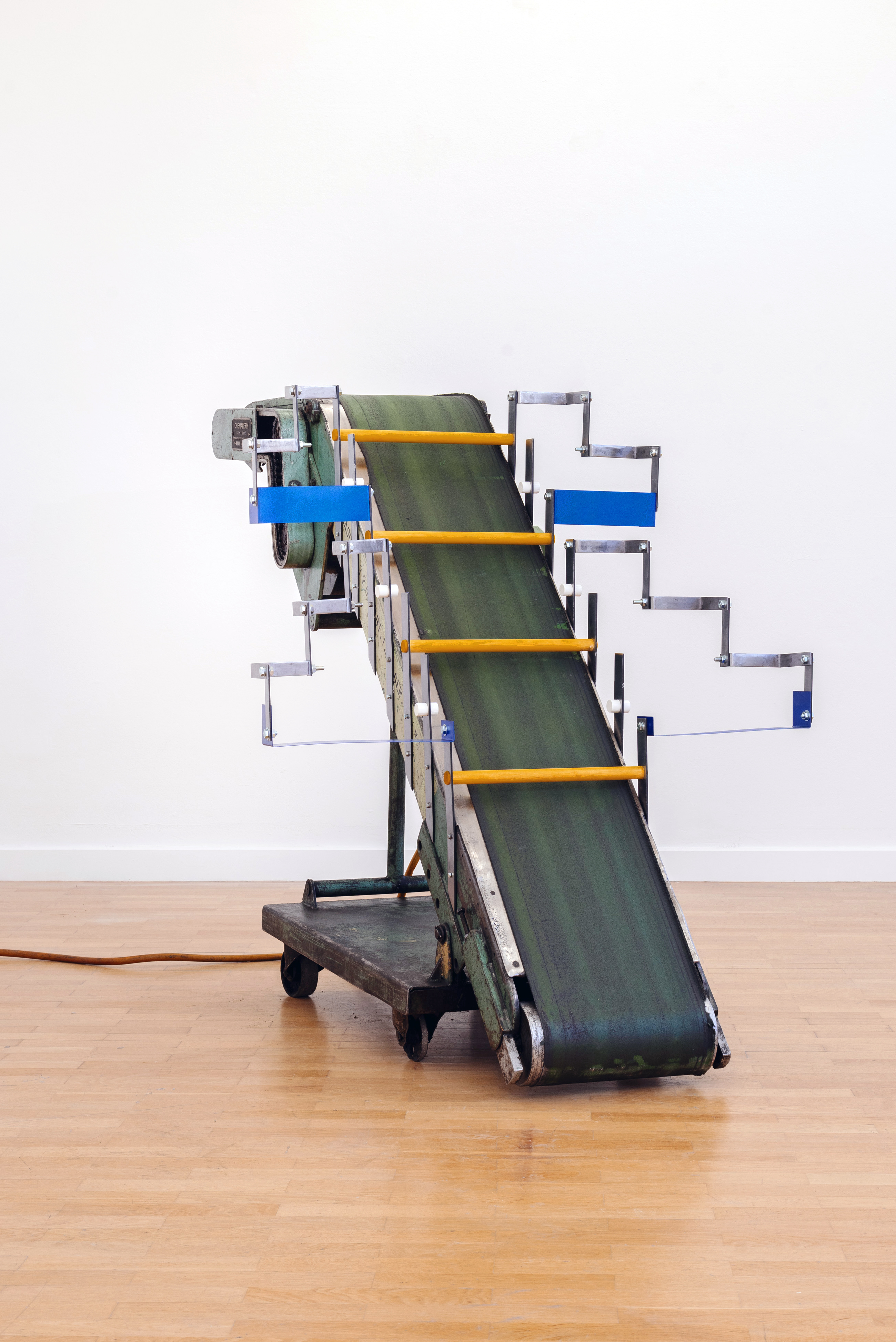 Skulptur aus einem gebrauchten Förderband auf Rädern, ergänzt durch Metallarme und farbige Querleisten. Das Objekt steht vor einer weißen Wand auf einem Holzboden.