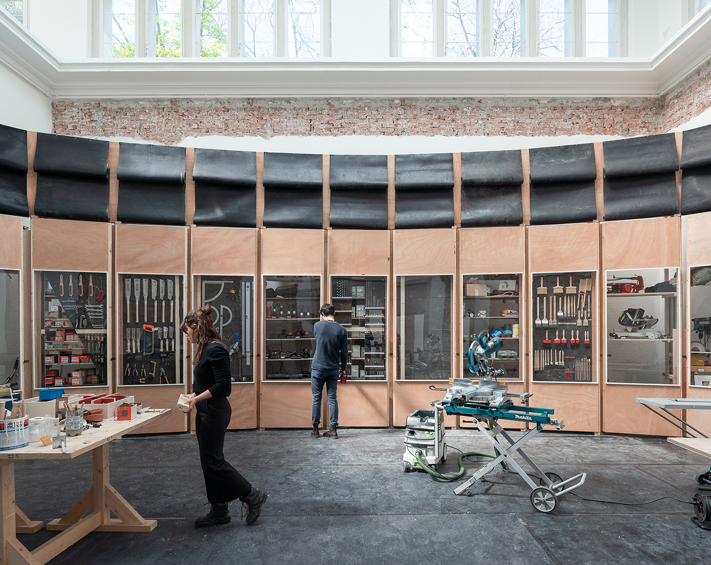 Werkstatt mit hohen Fenstern, Werkzeugwänden und zwei Personen, die an Arbeitsmaterialien und Geräten beschäftigt sind. Im Raum stehen Arbeitstische, Sägen und Maschinen.