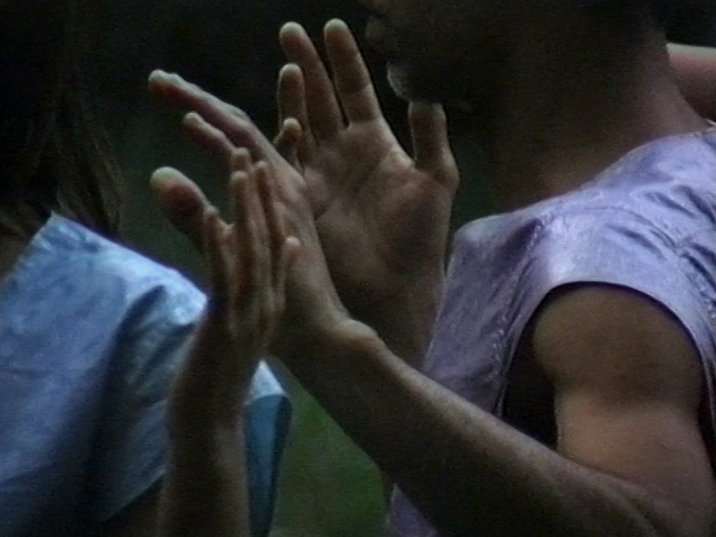 Nahaufnahme zweier Personen, deren Hände sich einander zuwenden, als würden sie eine Bewegung oder Geste ausführen. Sie tragen glänzende Oberteile in Blau- und Lilatönen.