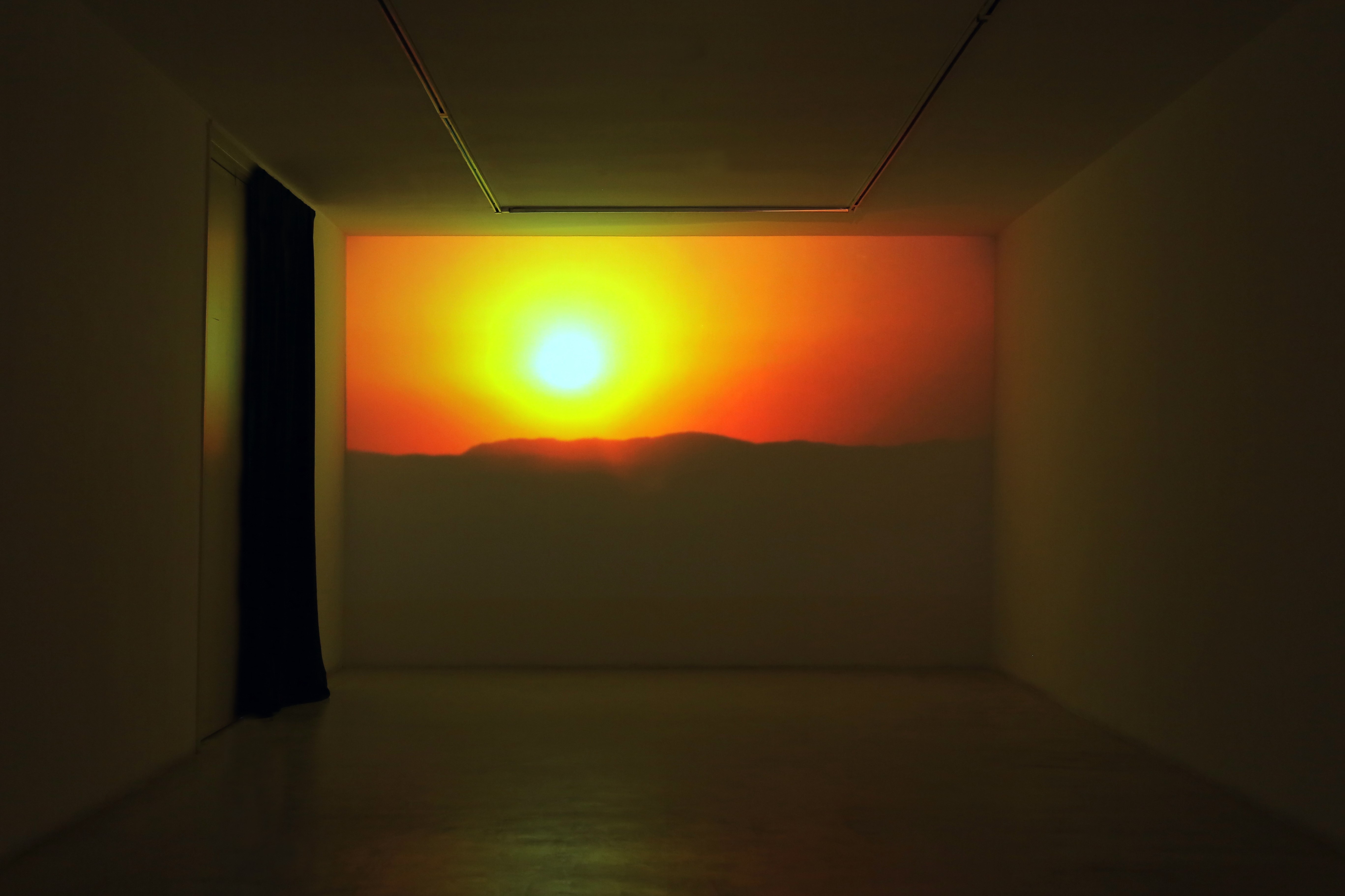 Darkened exhibition room with a large-scale light projection on the far wall, depicting a low sun above a dark horizon line in shades of yellow and orange.