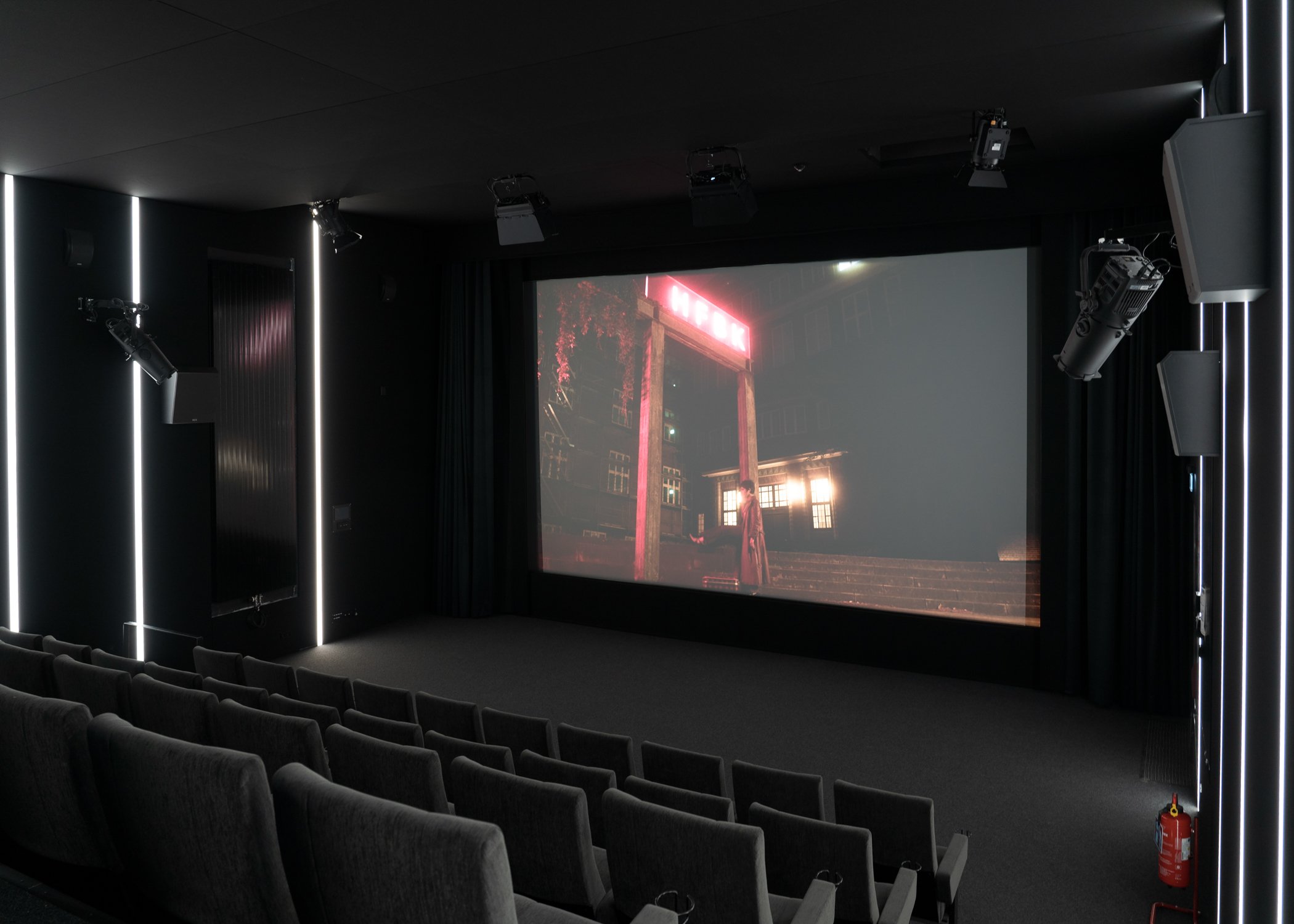 Dark cinema hall with rows of upholstered seats and a large screen displaying a film scene; technical lighting and speakers are mounted on the ceiling and walls.